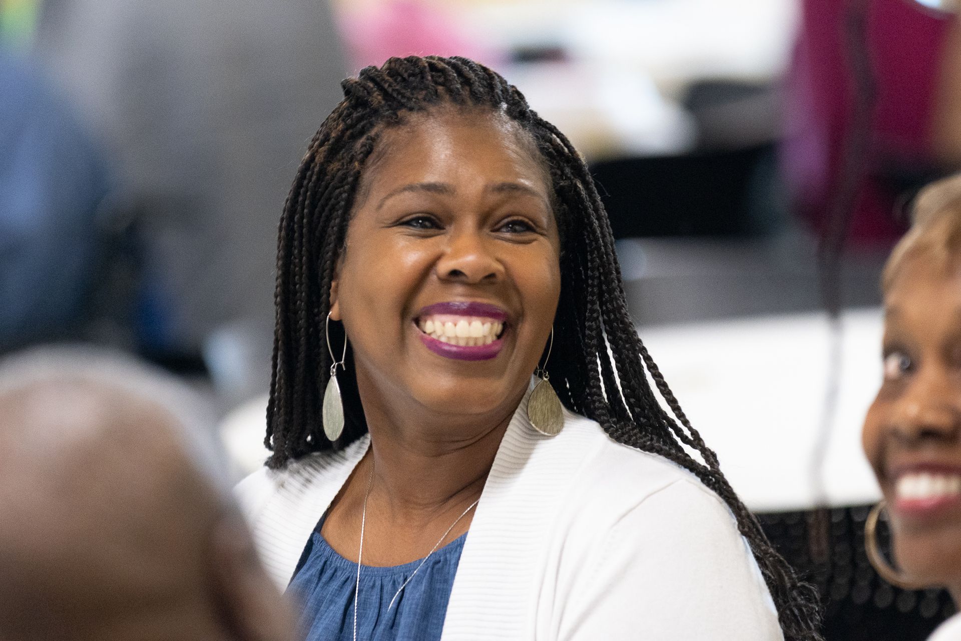 Woman with long braided hair, smiling and looking to the side, wearing a white cardigan and blue top.