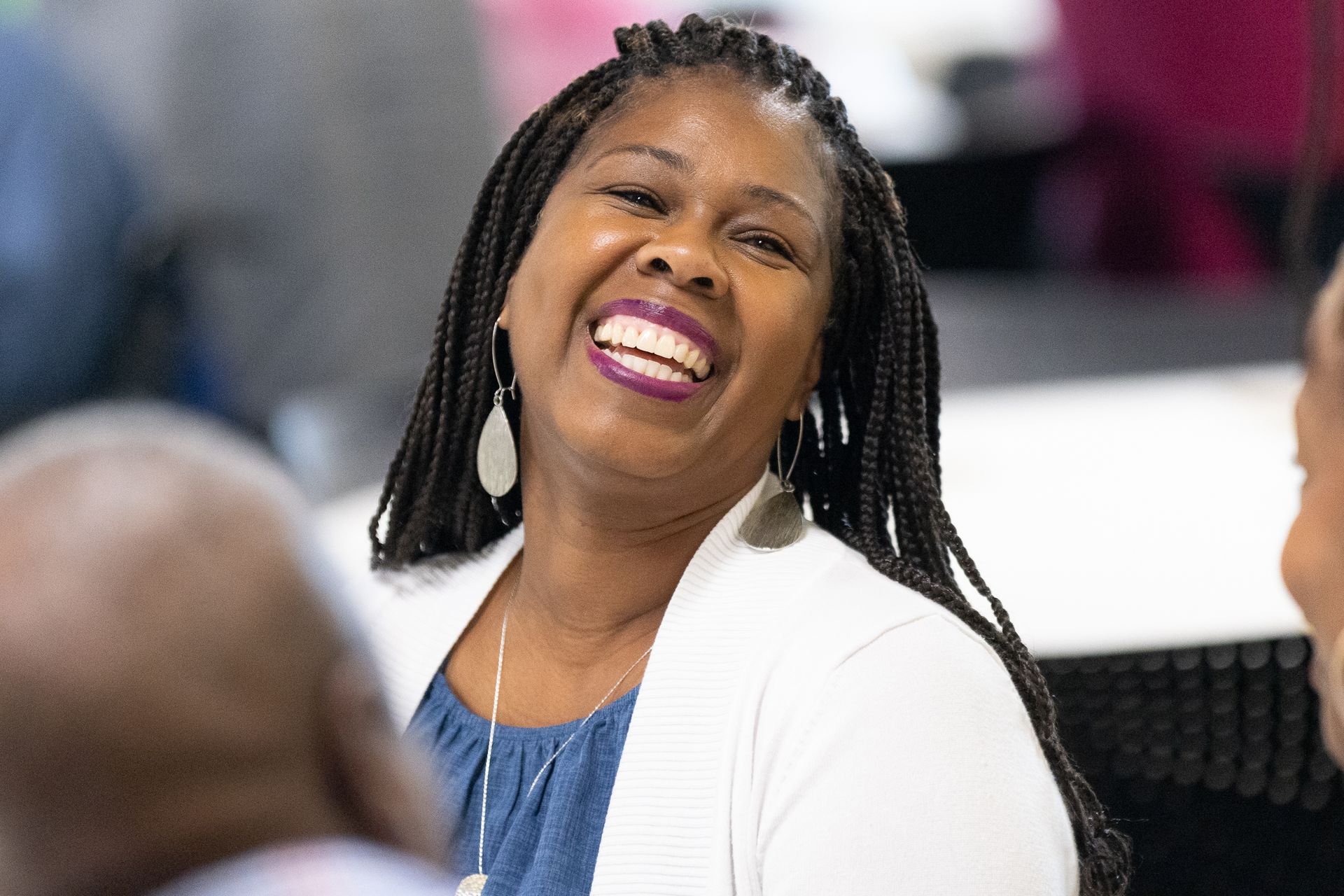 Woman smiling, wearing a white cardigan and blue shirt, with braided hair, in a room with others.