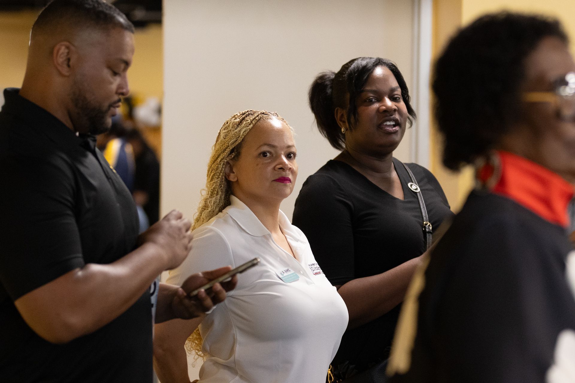 People in a hallway: one uses a phone, a woman with blonde hair, a woman talking.