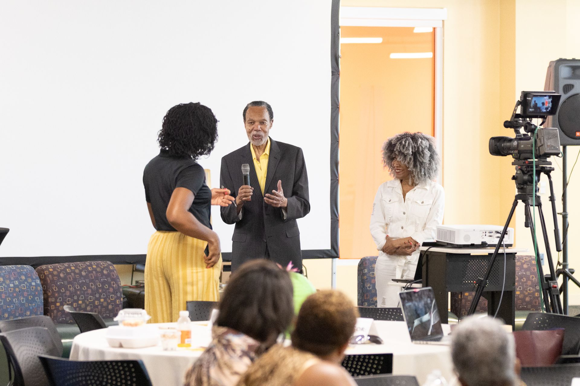 Three people on stage, speaking at an event, with a projection screen behind them.