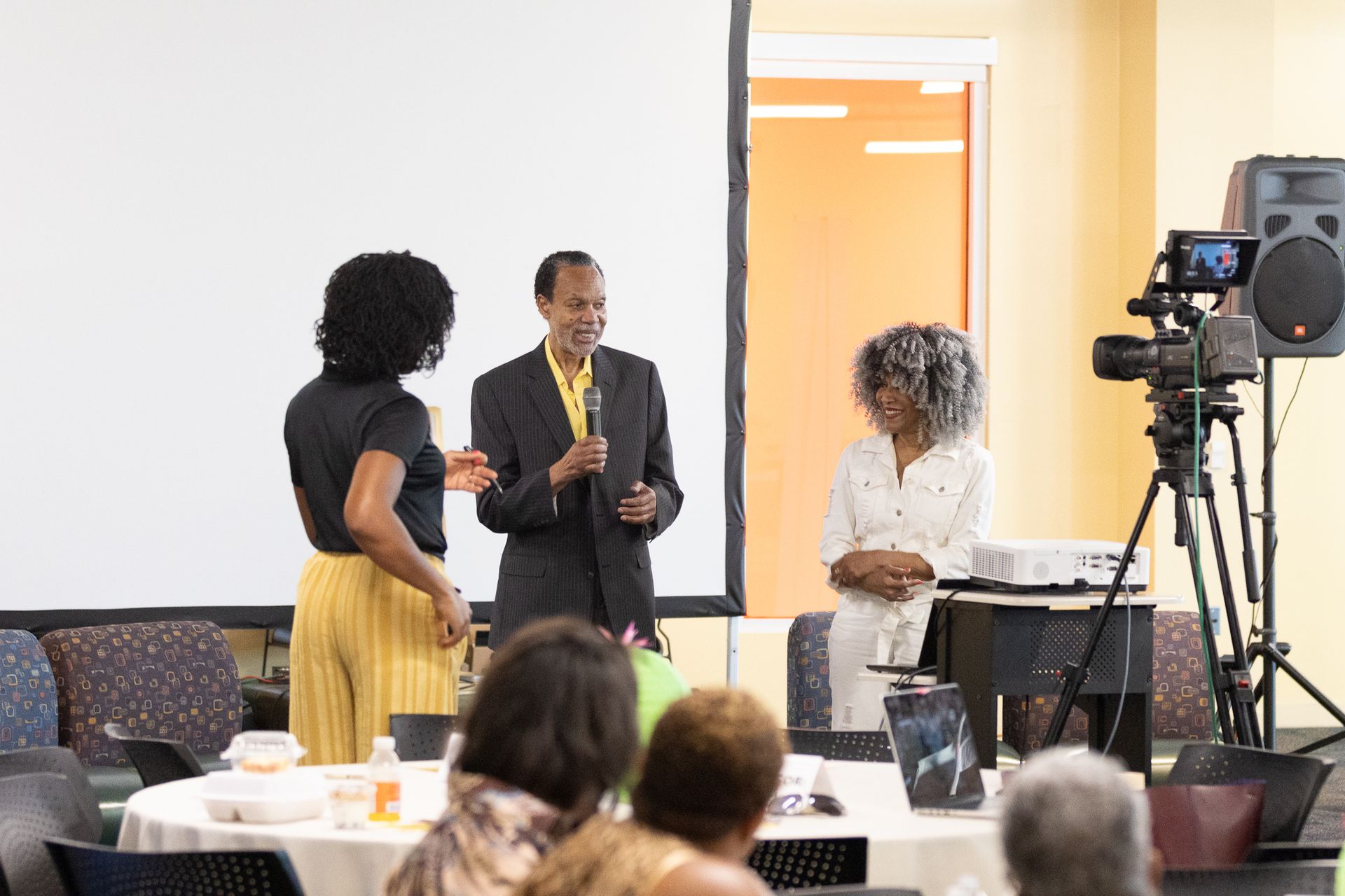 Three people on a stage with a microphone. One holds the microphone, while the others stand beside him. A camera films them.