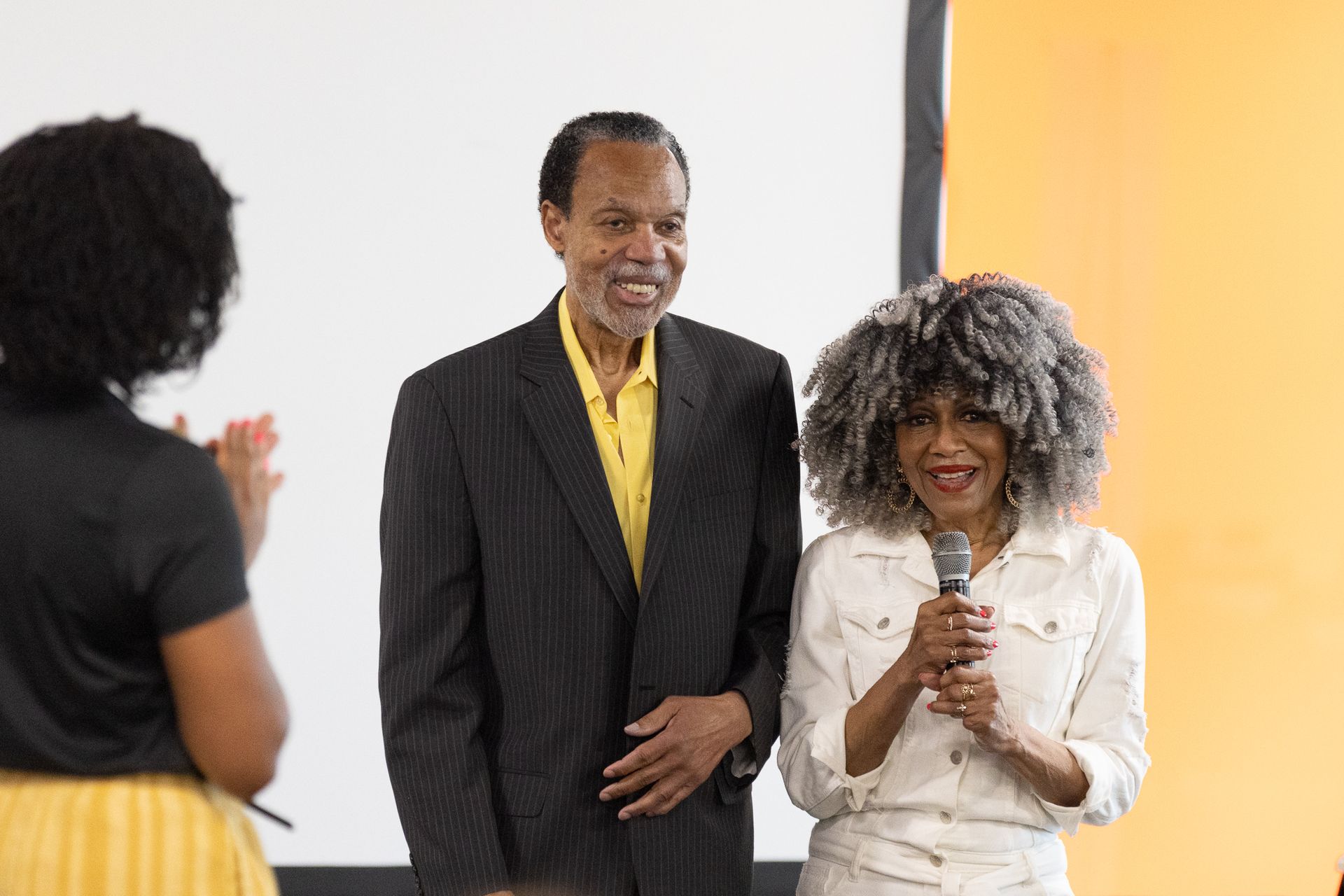 A man and woman standing, smiling, with a woman clapping. Behind them is a screen and an orange background.