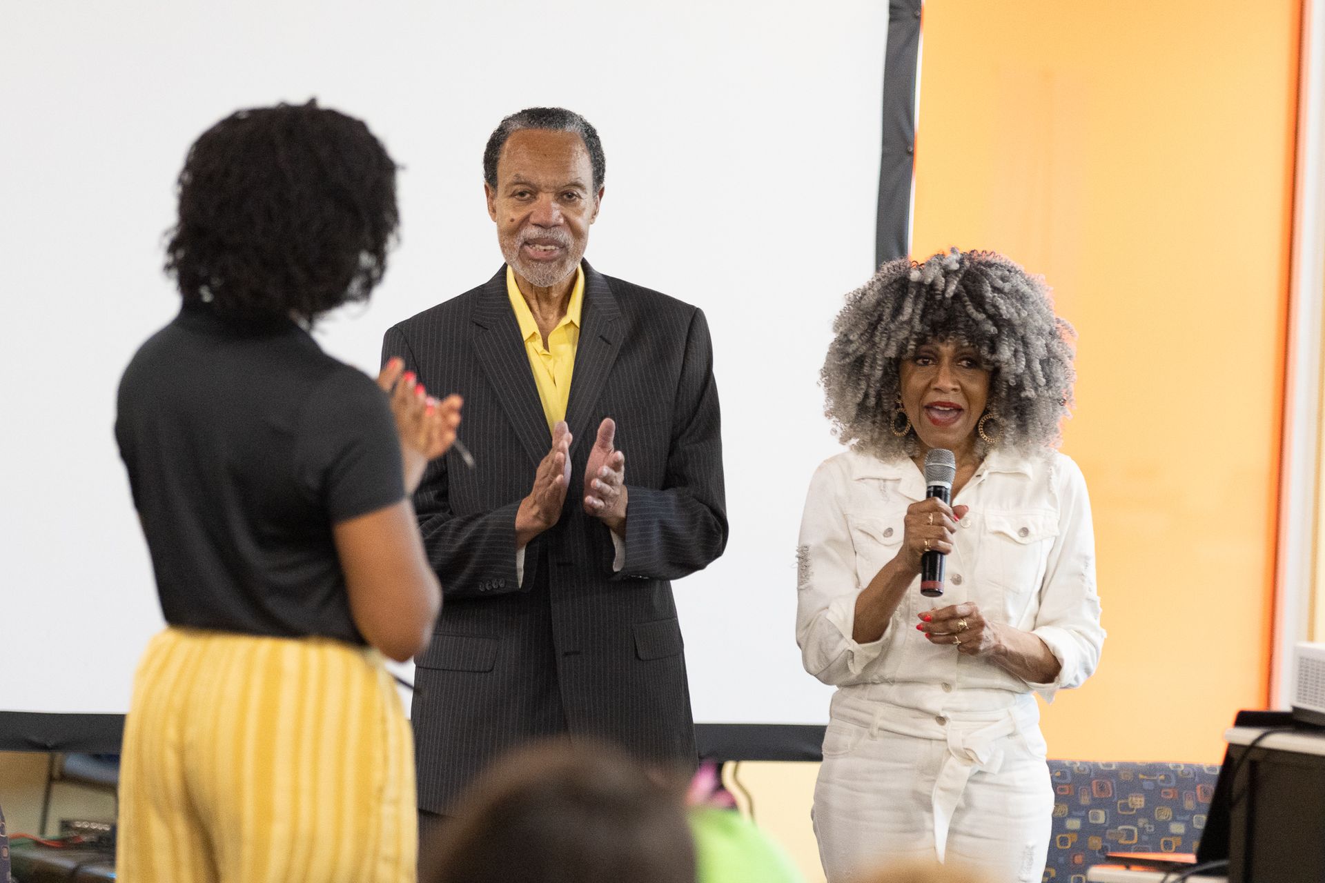 People on a stage: one woman speaking into a microphone, another clapping, and a man clapping.