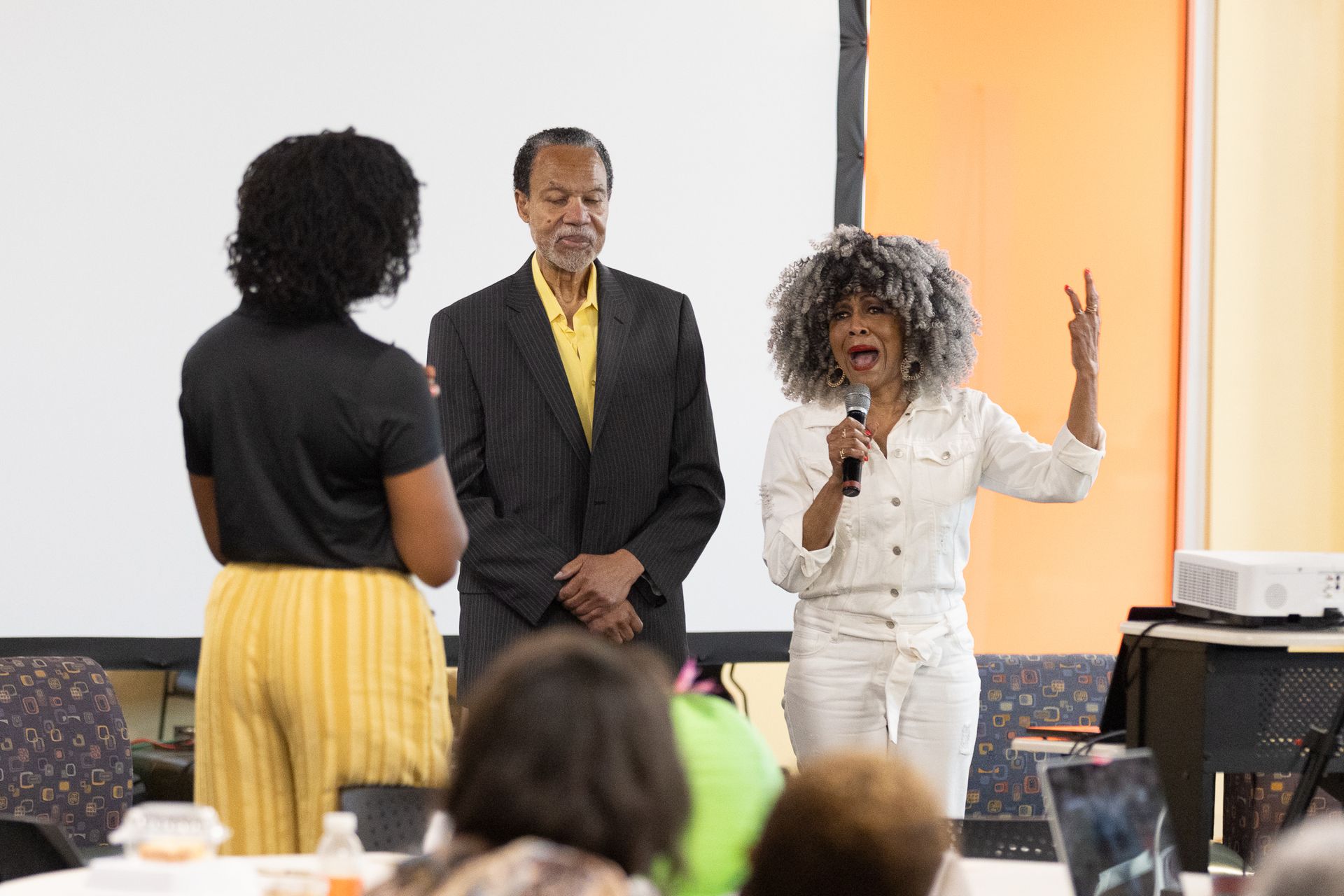 Woman speaking, gesturing, holding microphone. Two people stand near her, white wall and orange backdrop.