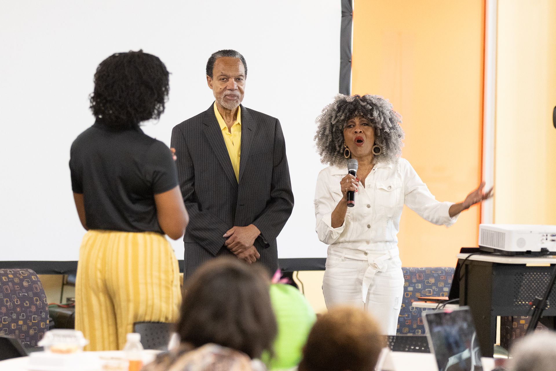 Woman speaks into microphone, gesturing; two others stand nearby in a room with a projector screen.