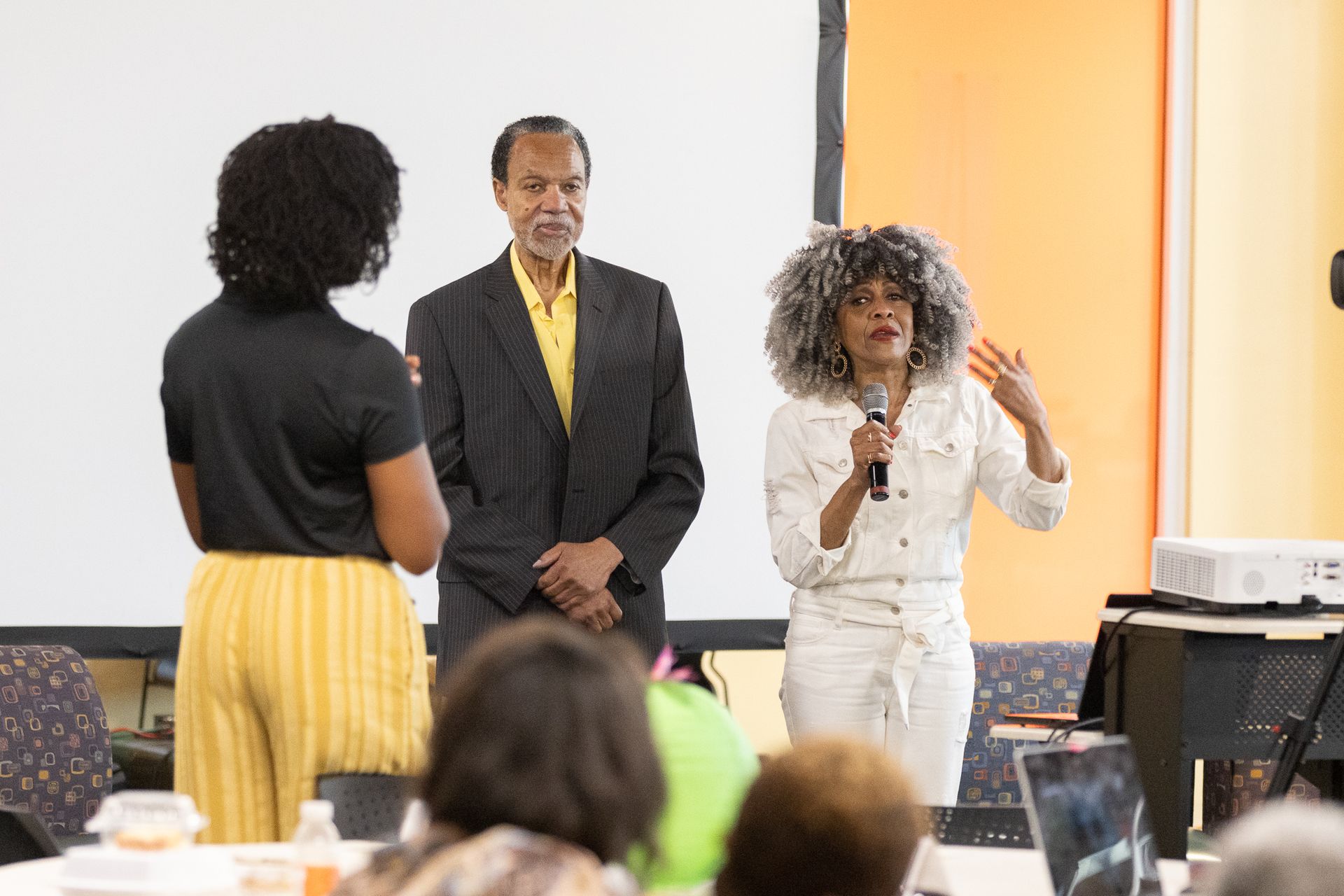 Woman speaking with a microphone, two others stand by a screen in a room with an audience.