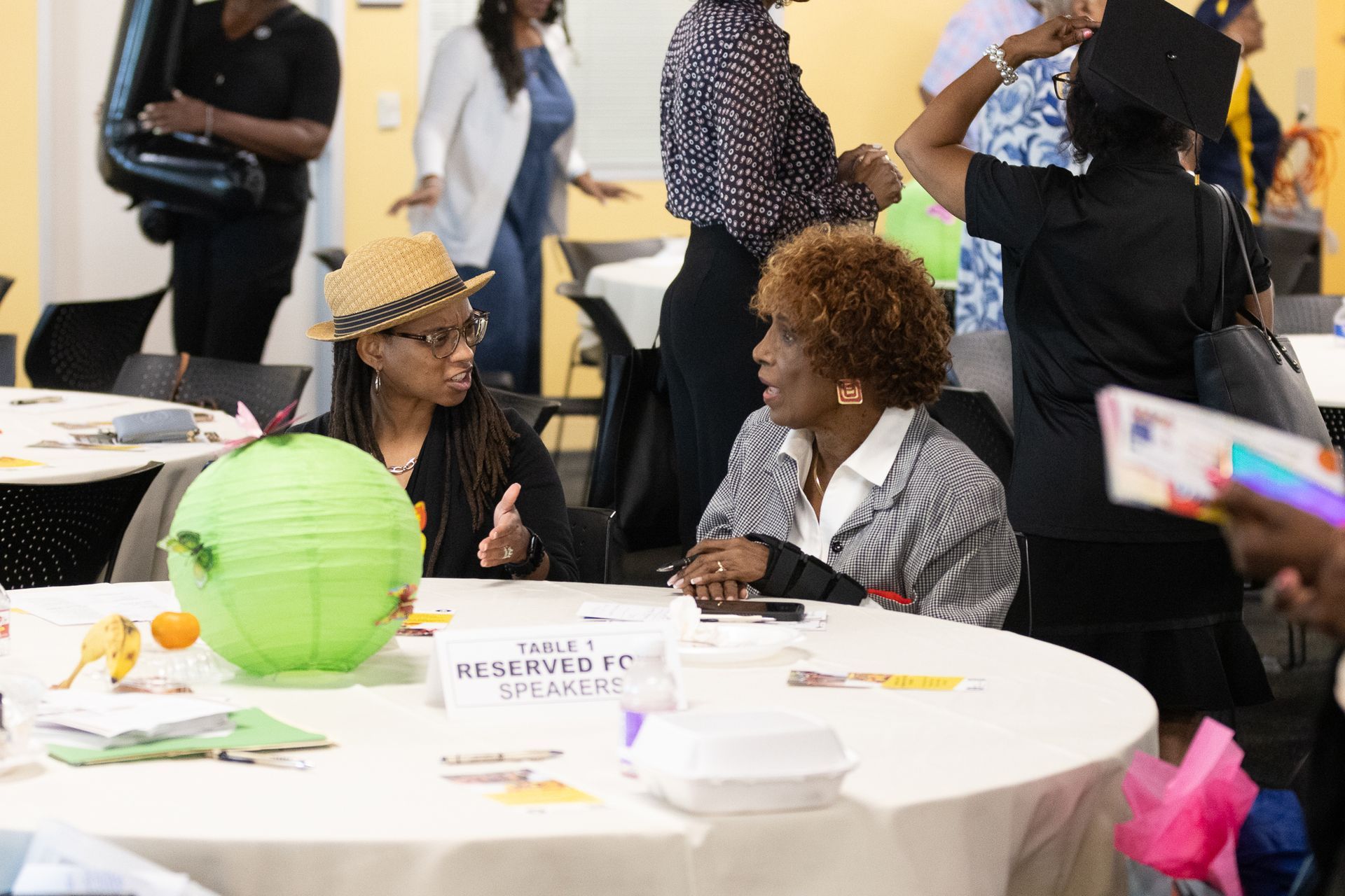 Two women converse at a round table; a straw hat and green paper lantern are on the table.
