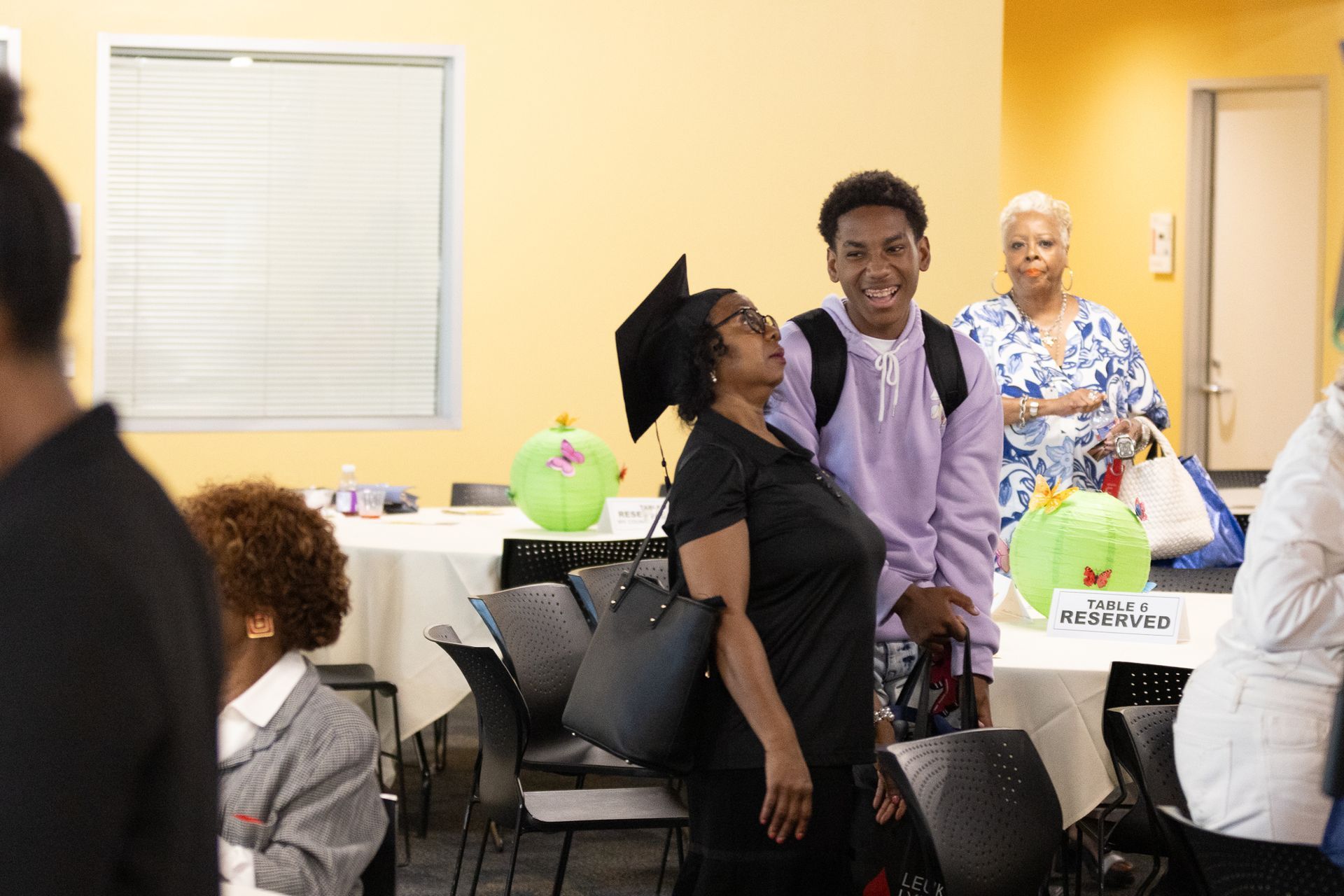 People at an event. A woman in a graduation cap looks up at a smiling young person. Tables with decorations in a yellow room.