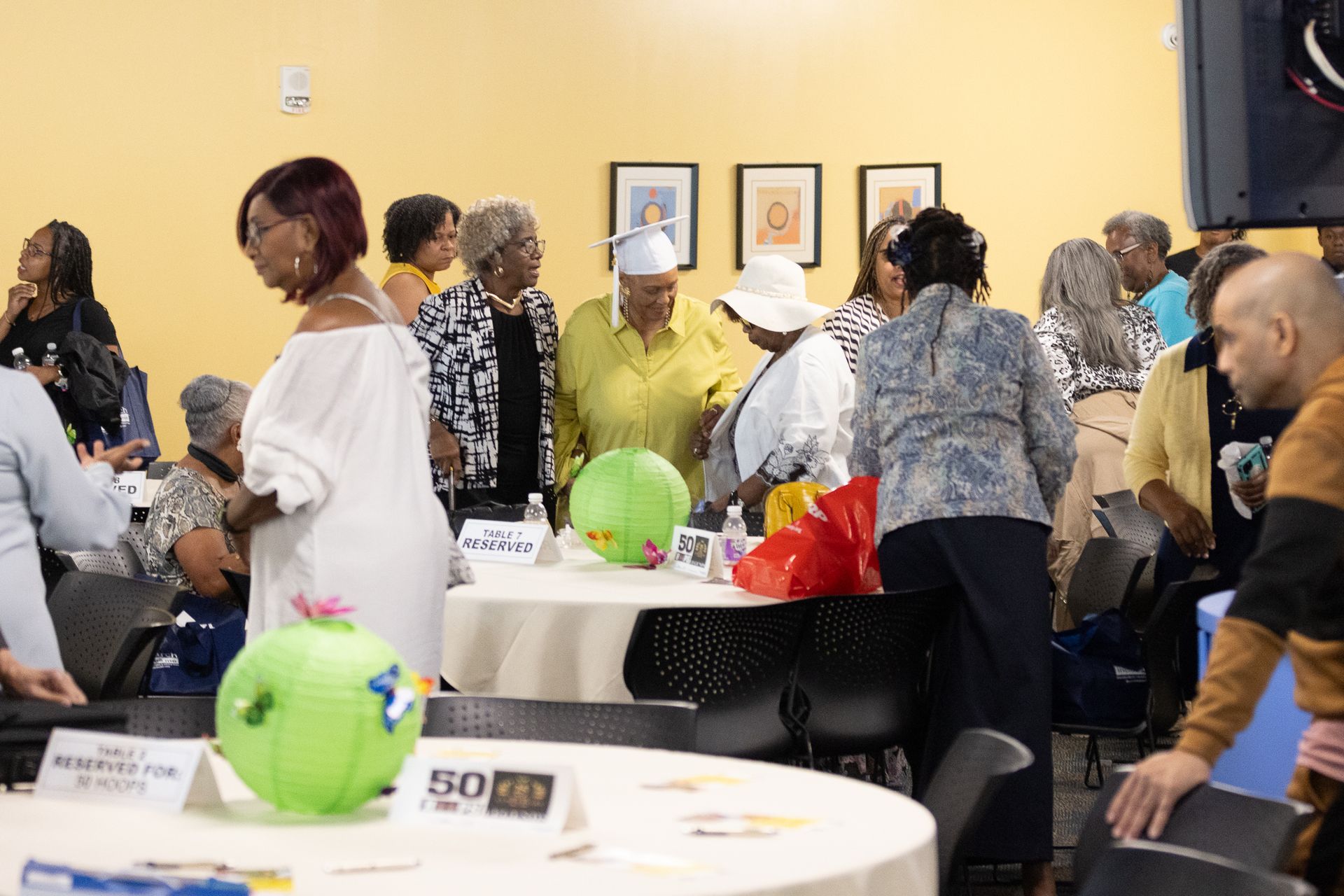 People gather around tables indoors; some are looking at items. Green and red decorations are on a table.