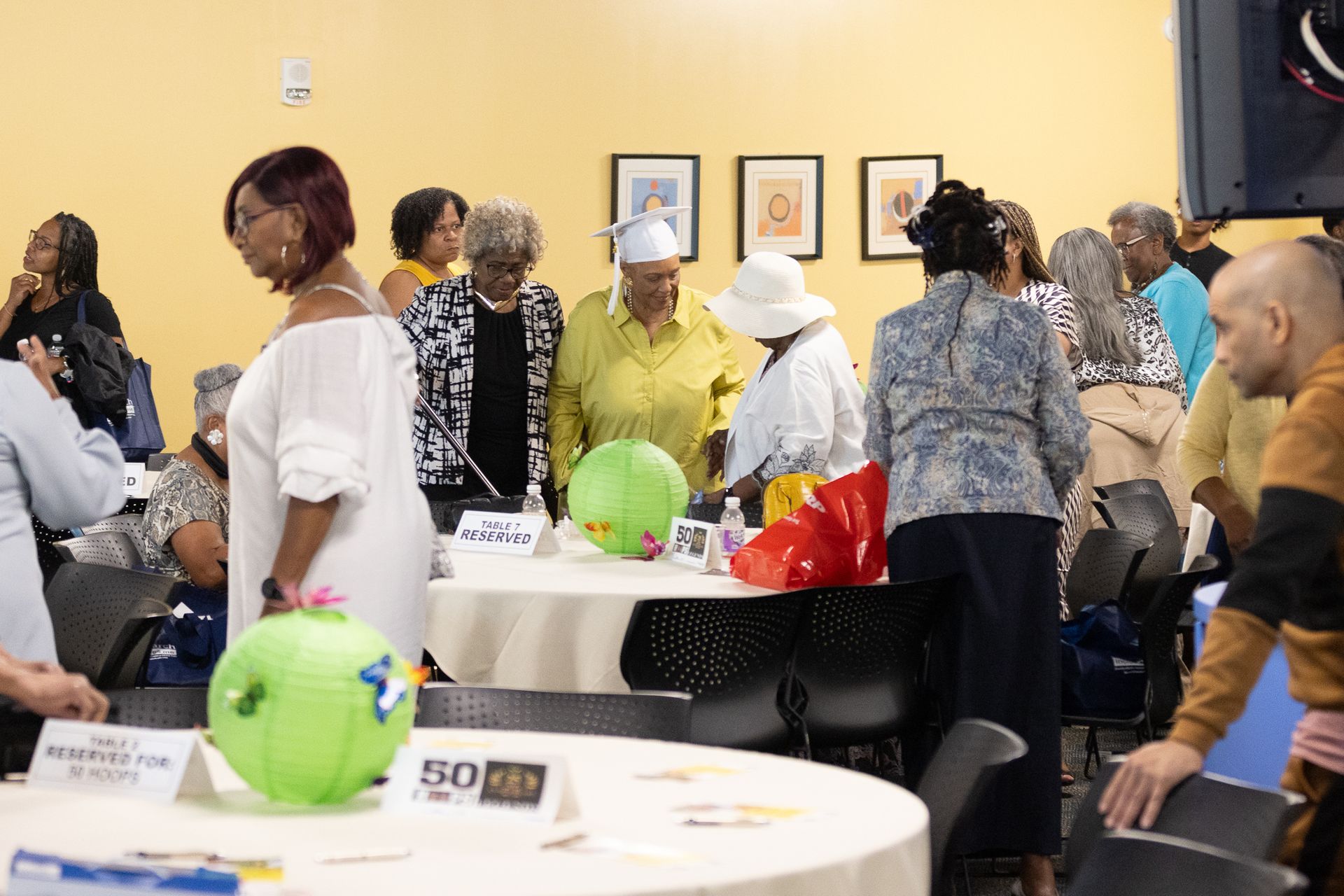 People gathered at tables, balloons, and framed artwork in a yellow-walled room.
