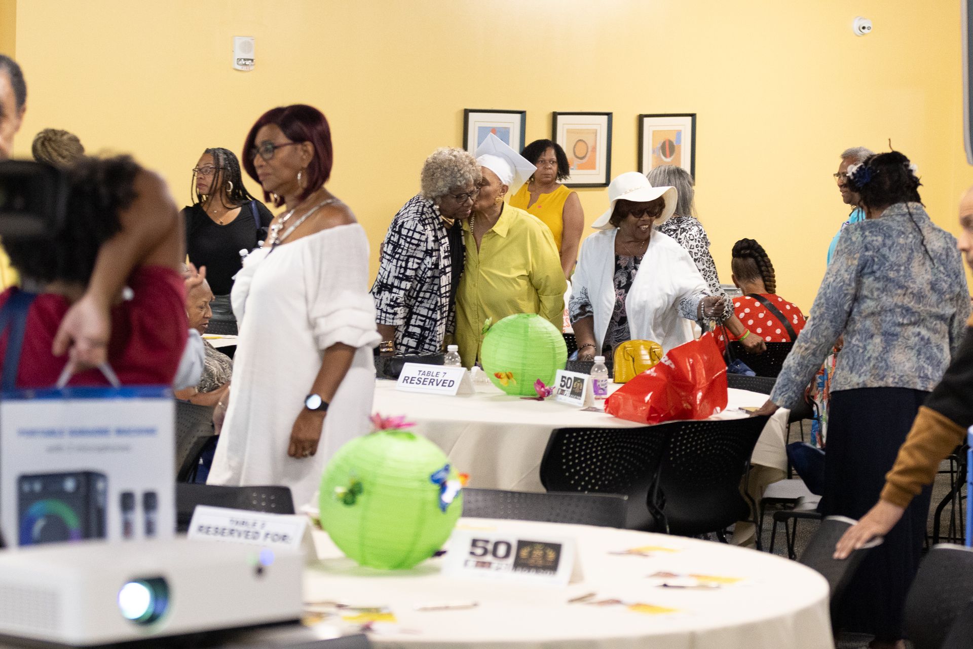 People mingling in a brightly lit room with decorated tables; a projector sits in the foreground.