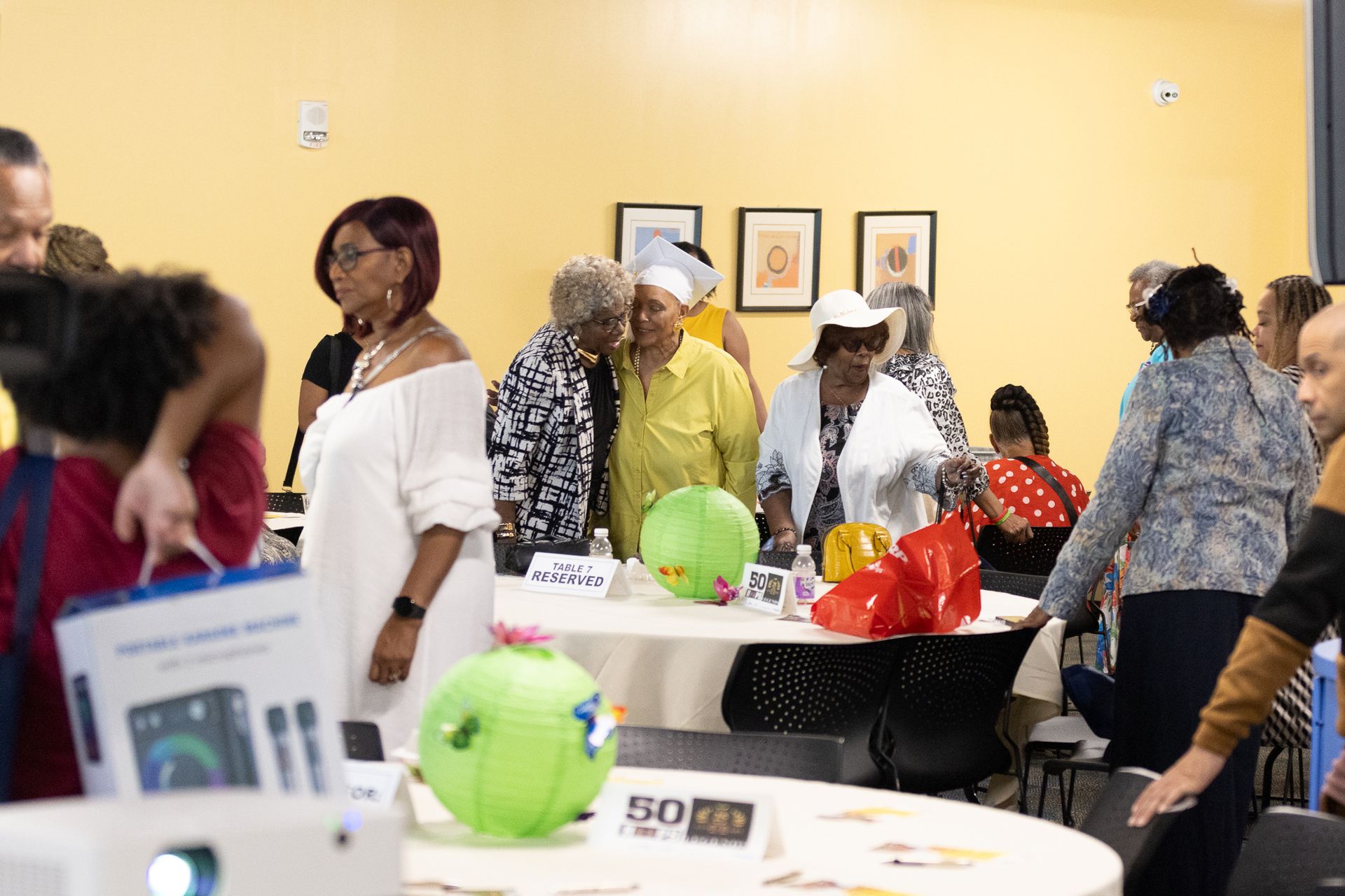 People gathered around tables, likely at an event. Some are looking at decorations, others are conversing.
