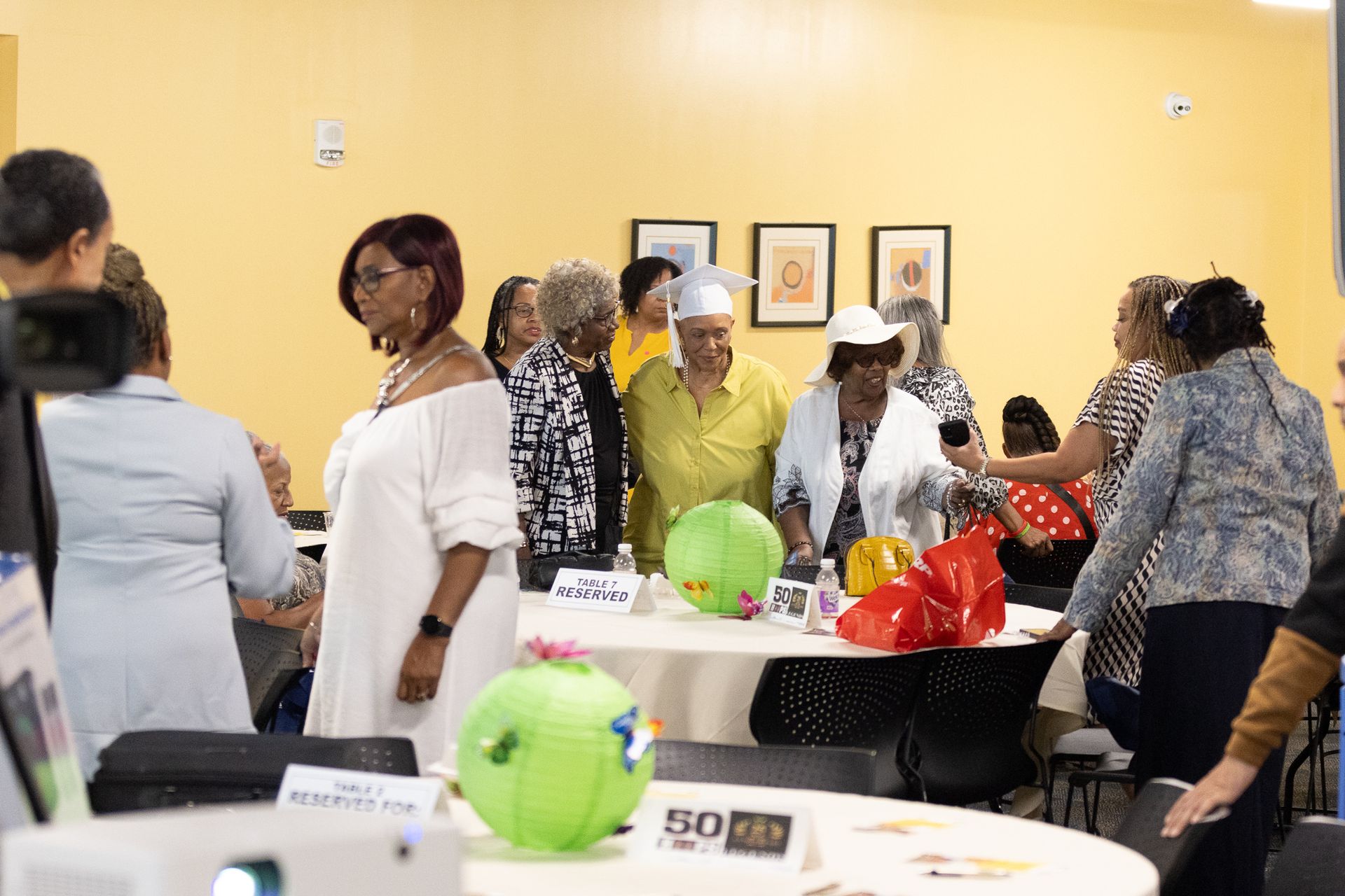 People gathered in a yellow-walled room, around tables. Some are looking at objects and others talking.