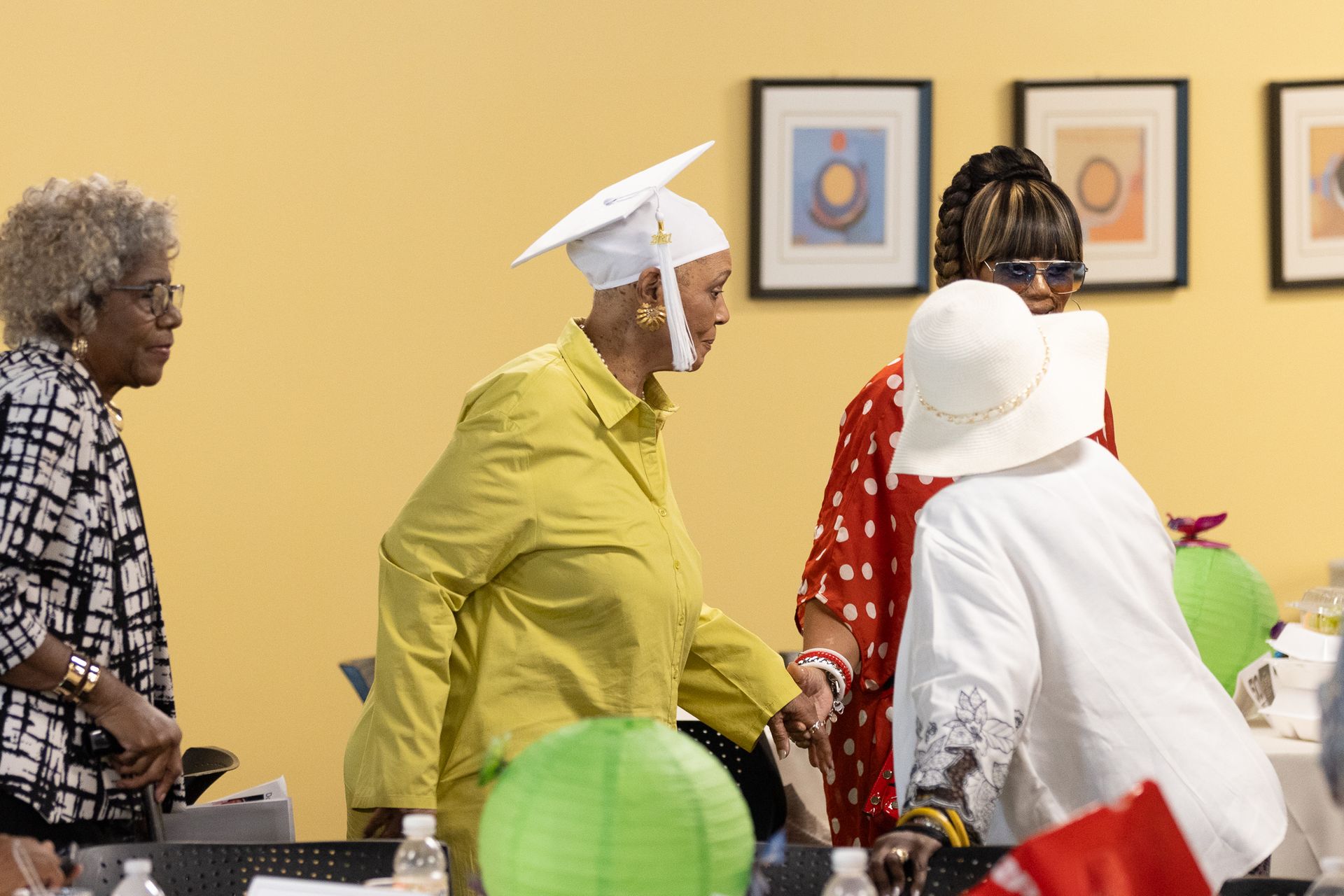 Group of people at a celebration, one in a yellow suit and graduation cap. Artwork on the wall in the background.