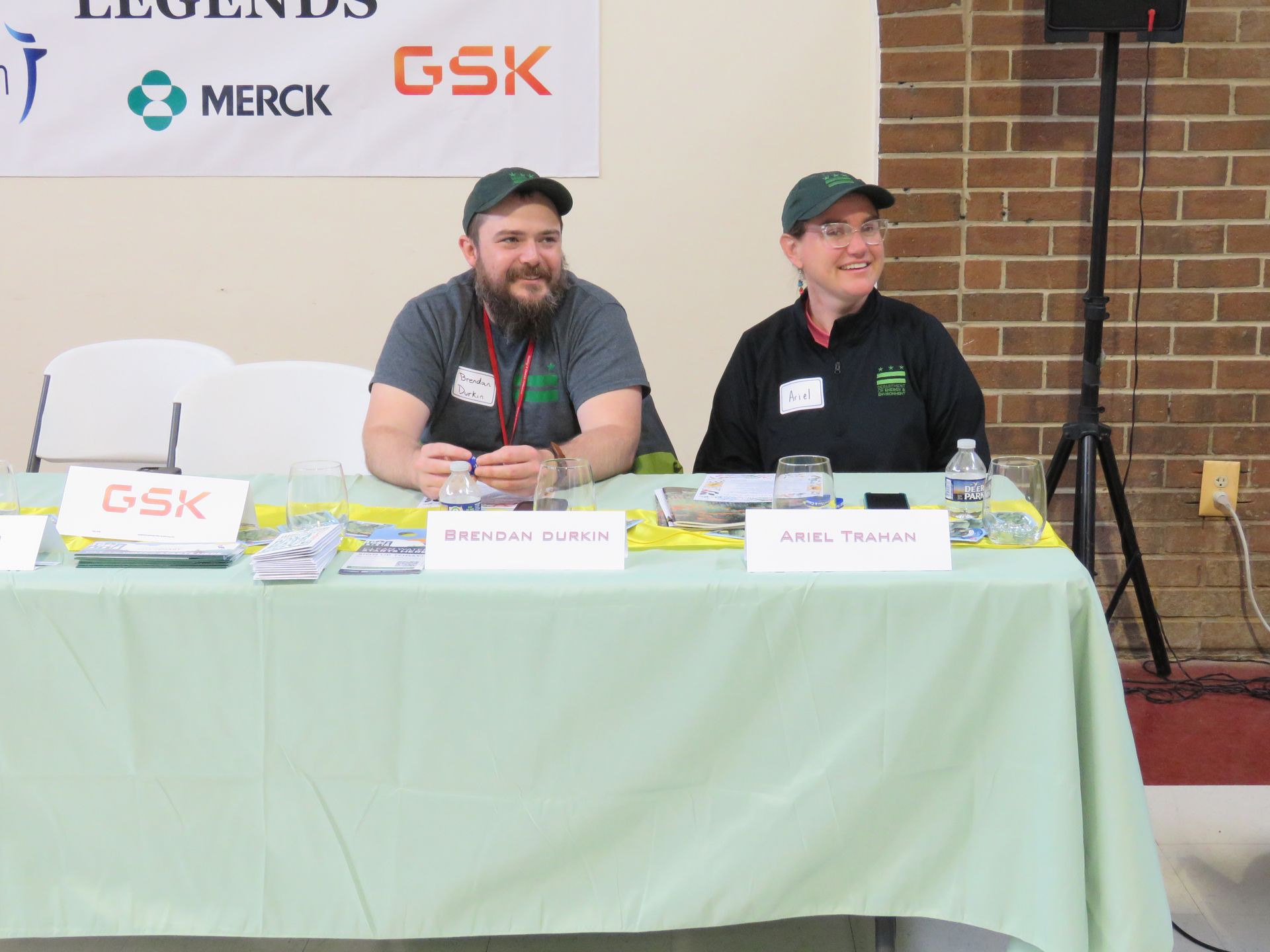 Two people sit behind a table with a GSK sign. They are wearing green shirts and smiling.