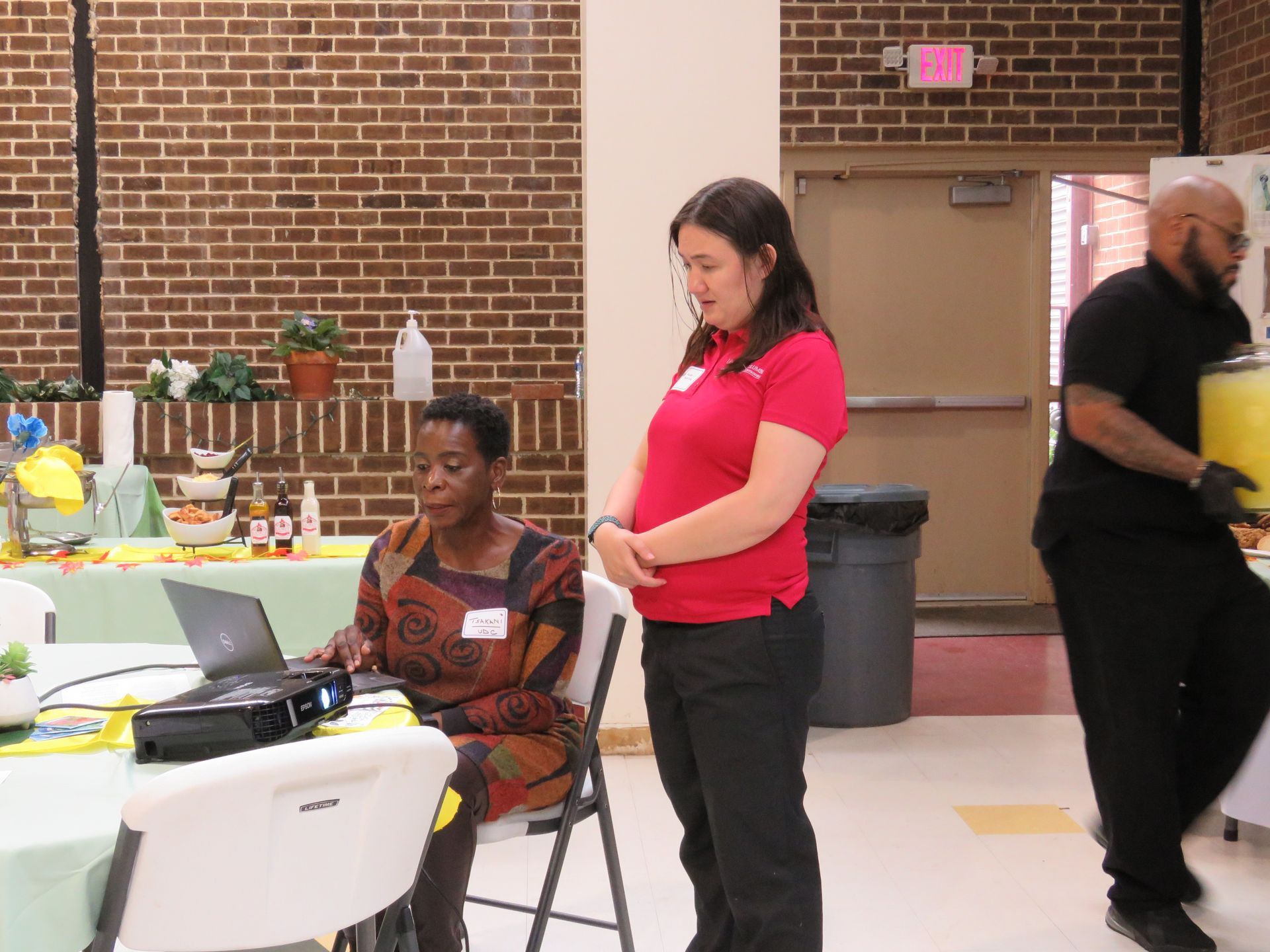 Woman working on laptop, two others stand nearby in a room with brick walls.