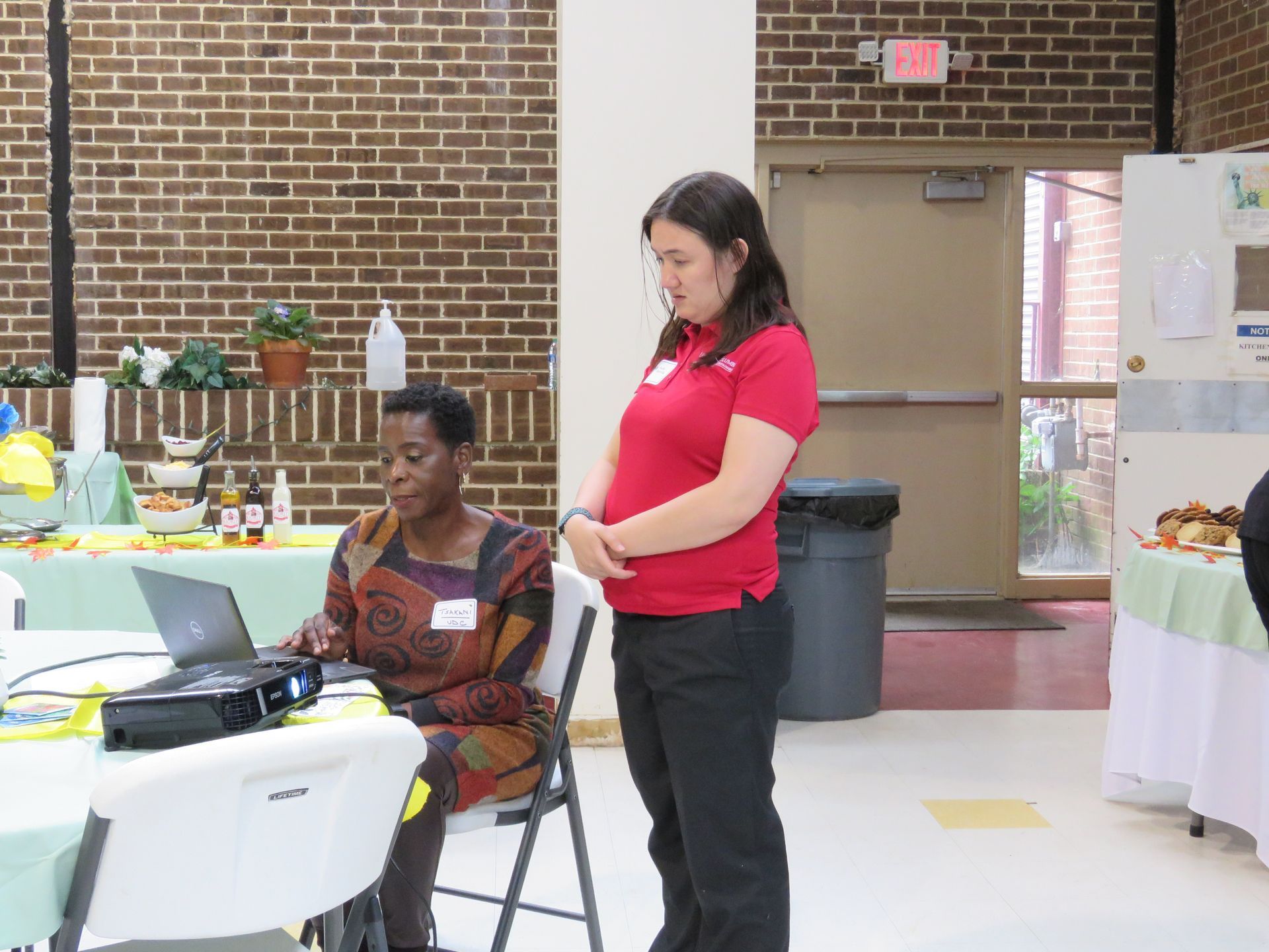 Woman seated at laptop, another standing beside. Brick wall and decorated tables in a room.
