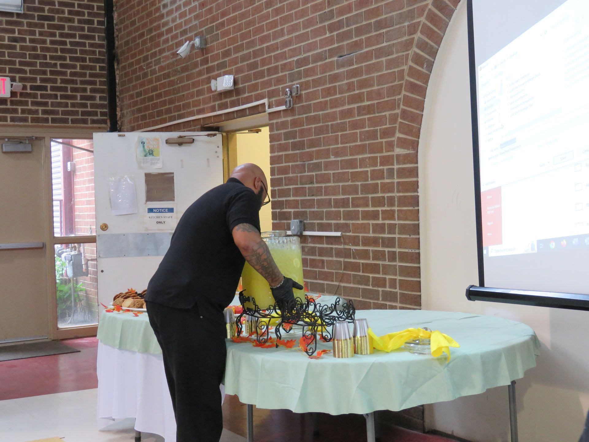Man in black shirt and mask, arranging items on a table with a pale green tablecloth in a room with a brick wall.