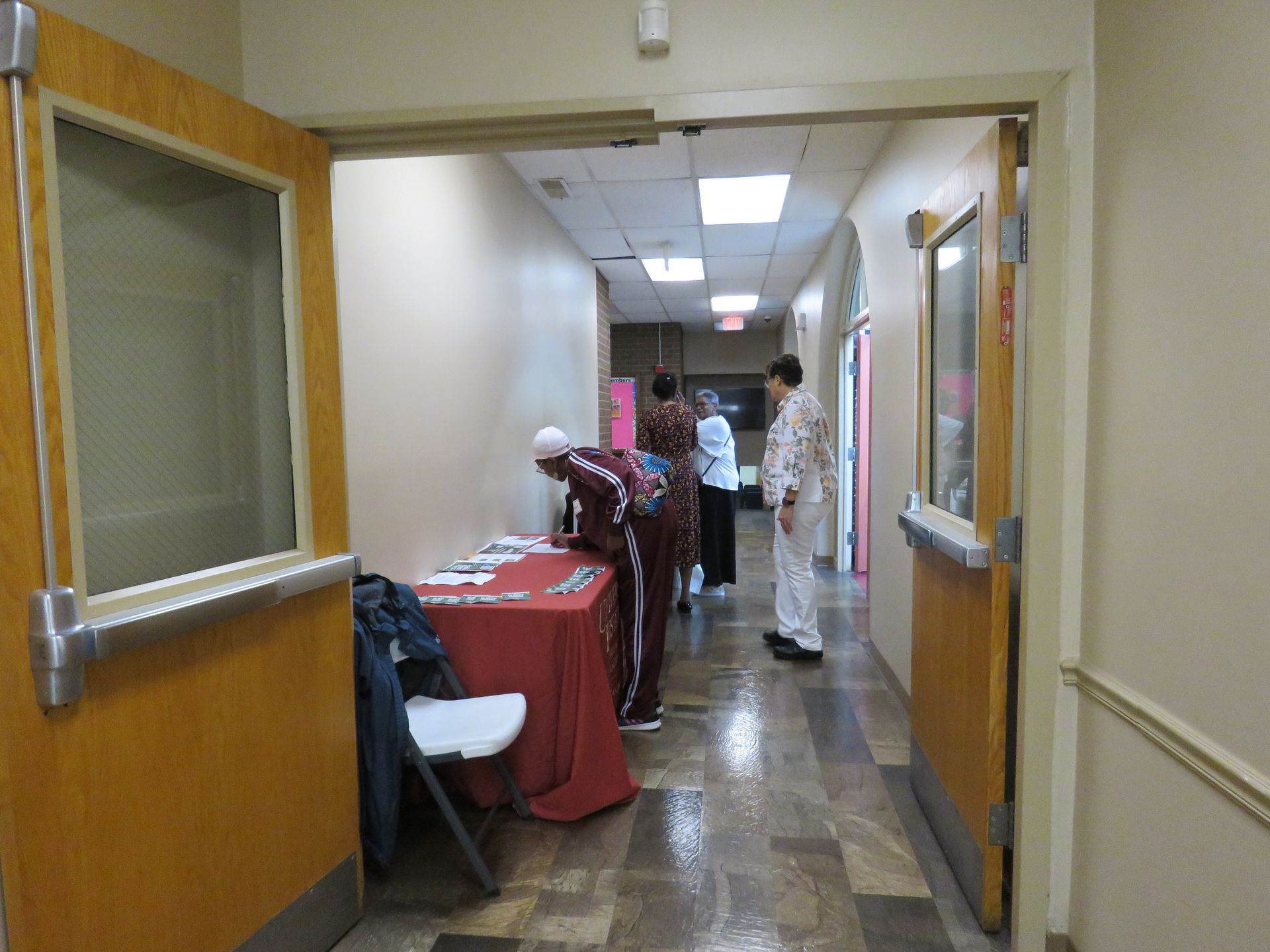 Hallway with people near a table, one in maroon and others in white and blue, and open doors on both sides.