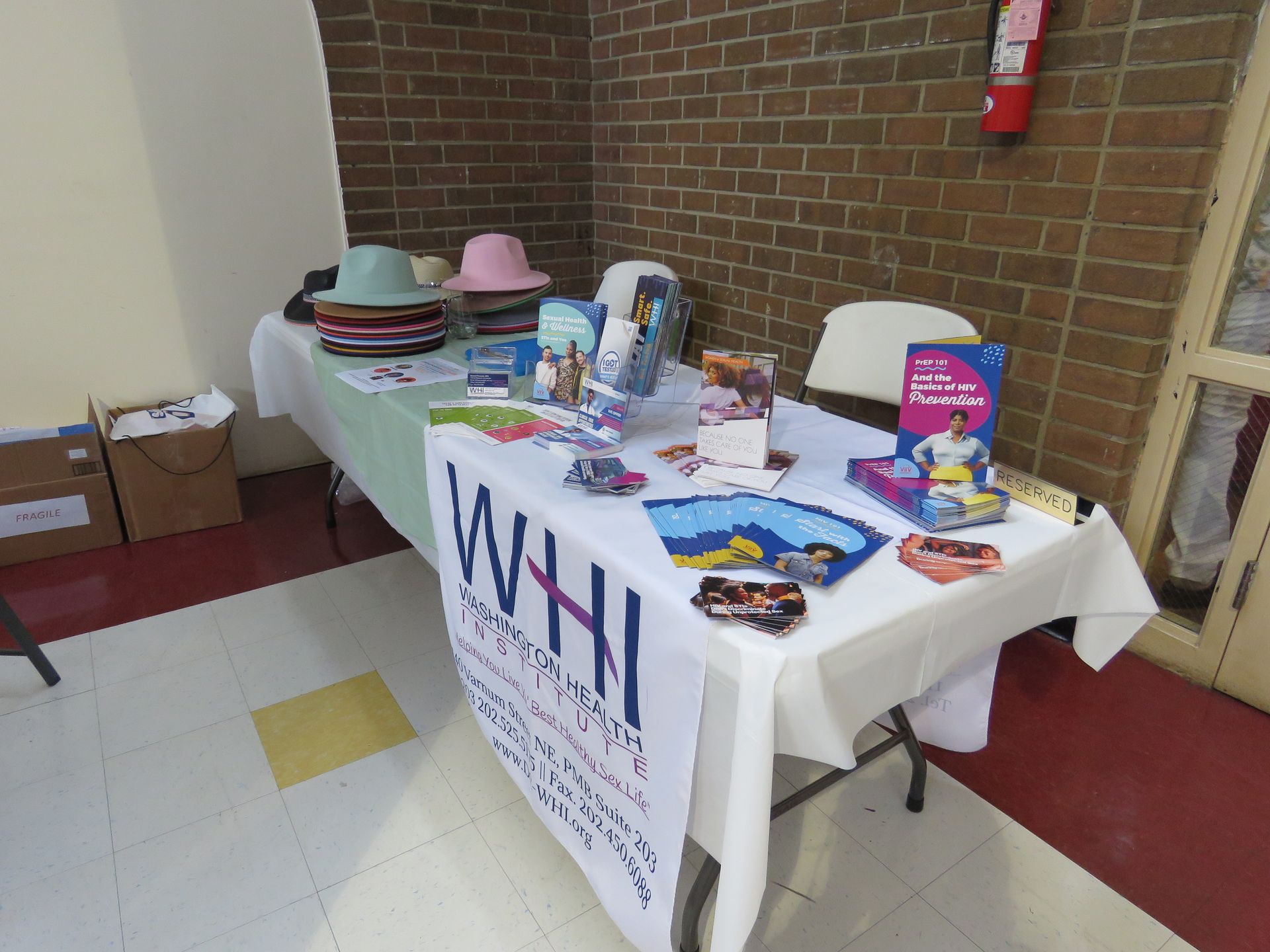 A table with hats, brochures, and a banner for a Women's Health Initiative event in a brick-walled room.