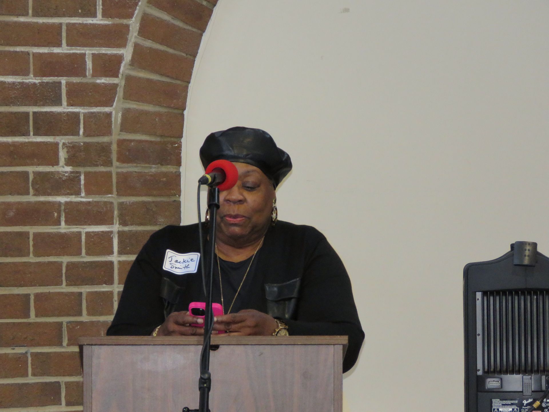 Woman speaking at a podium, holding a pink phone. Brick wall and arch background, interior setting.