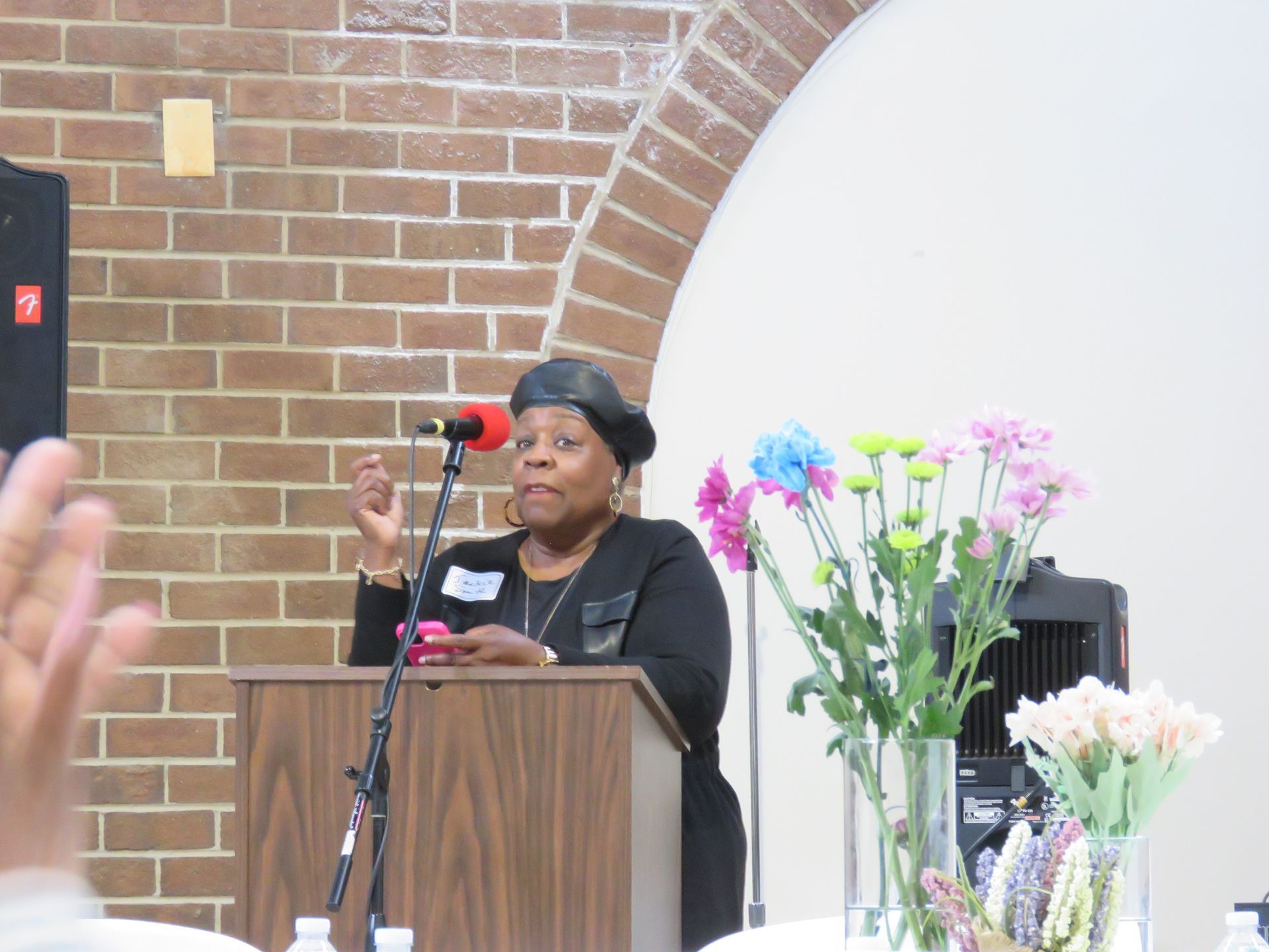 Woman speaking at a podium, gesturing with her hand, in front of a brick wall and floral arrangement.