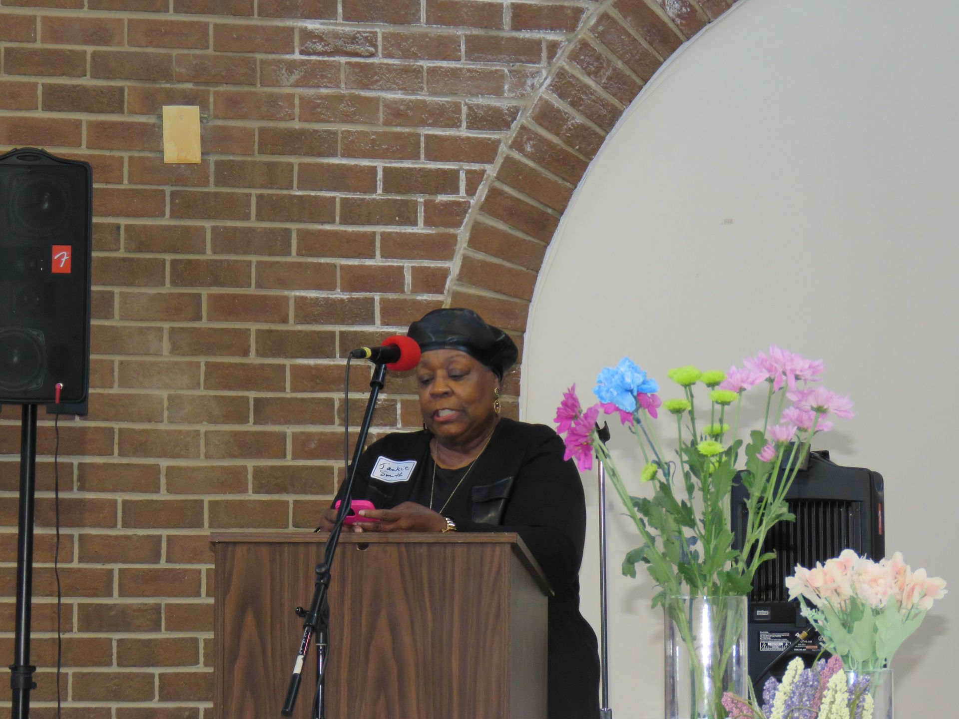 Woman speaking at a podium with a microphone. Brick wall backdrop, flowers on right.