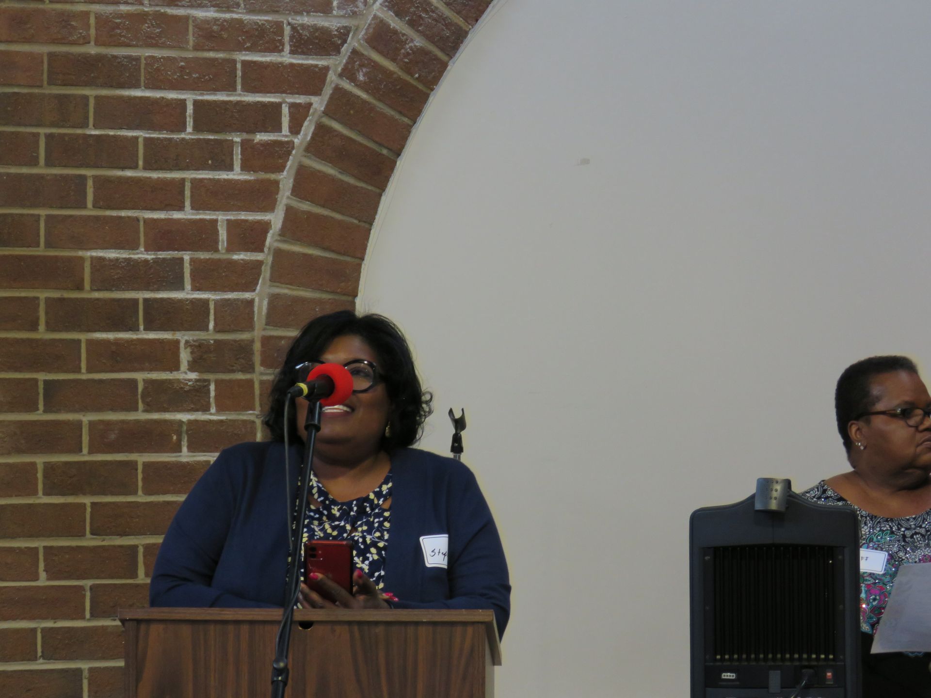 Woman speaking at a podium, brick wall background, another woman seated beside.