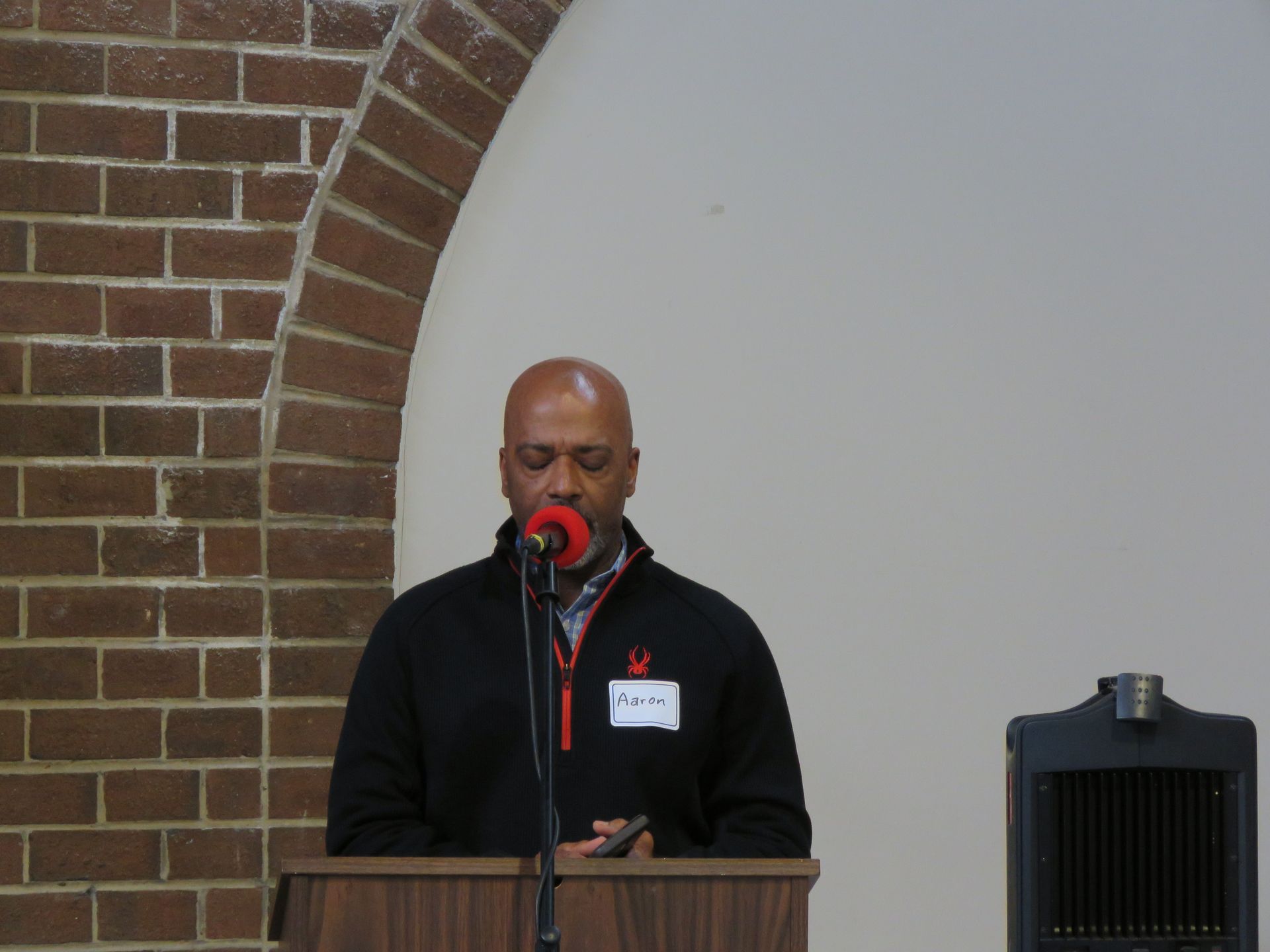 Man speaking at a podium with a brick archway backdrop and a black speaker.
