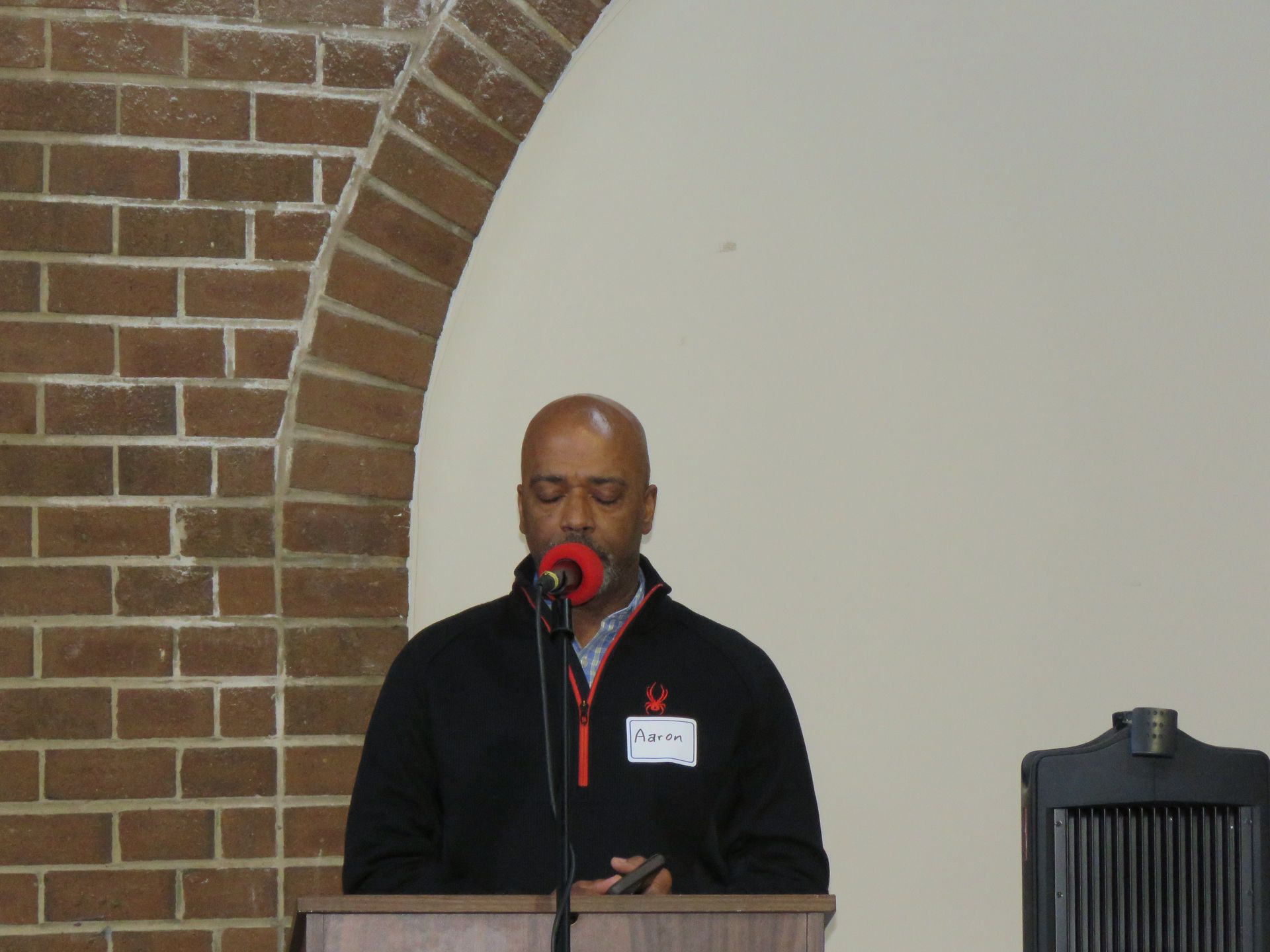 Man speaking at a podium, red microphone, brick archway, cream wall, and black speaker.