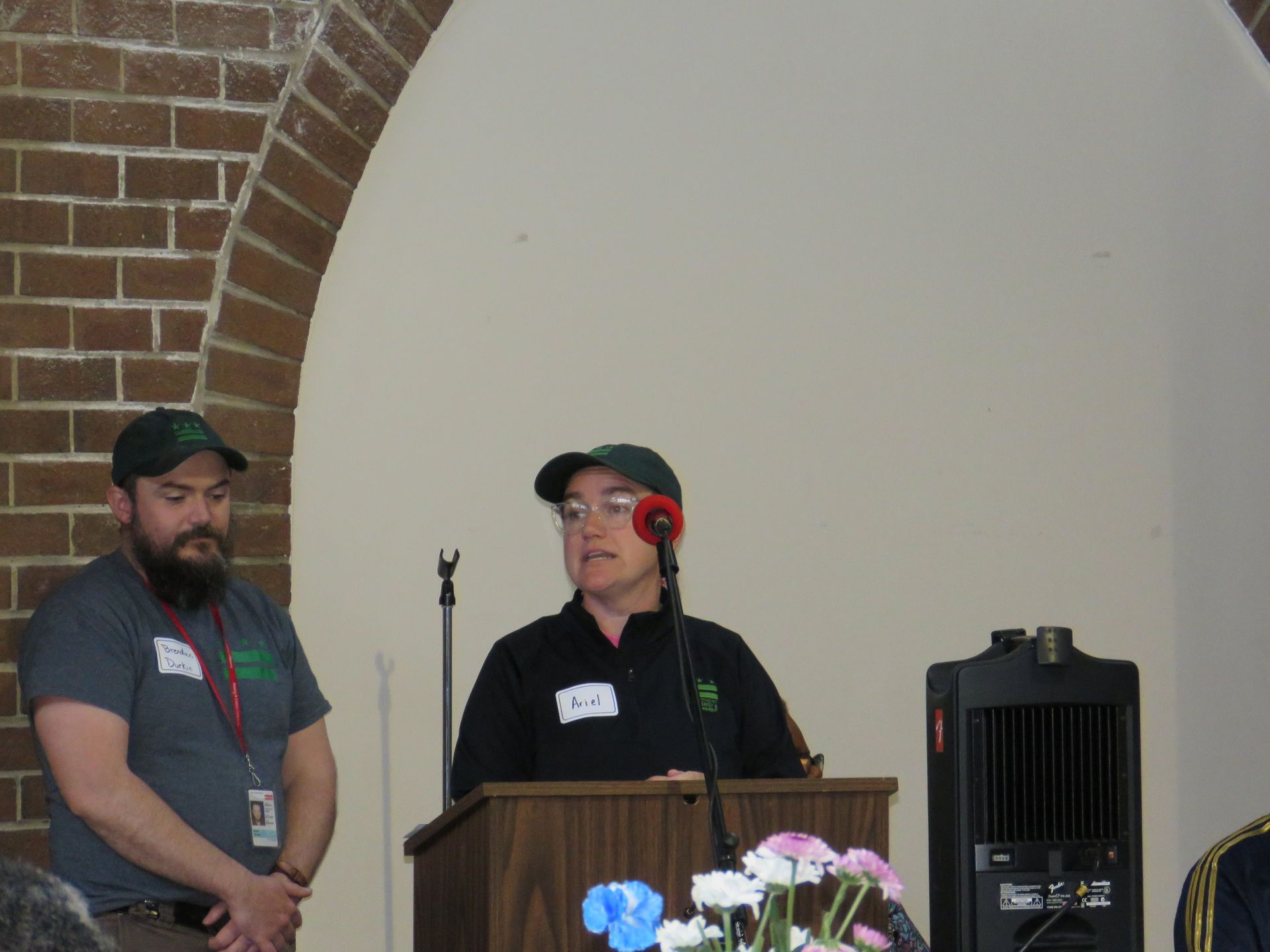 Two people at a podium with microphone. One speaks while another stands by, both wearing green hats.