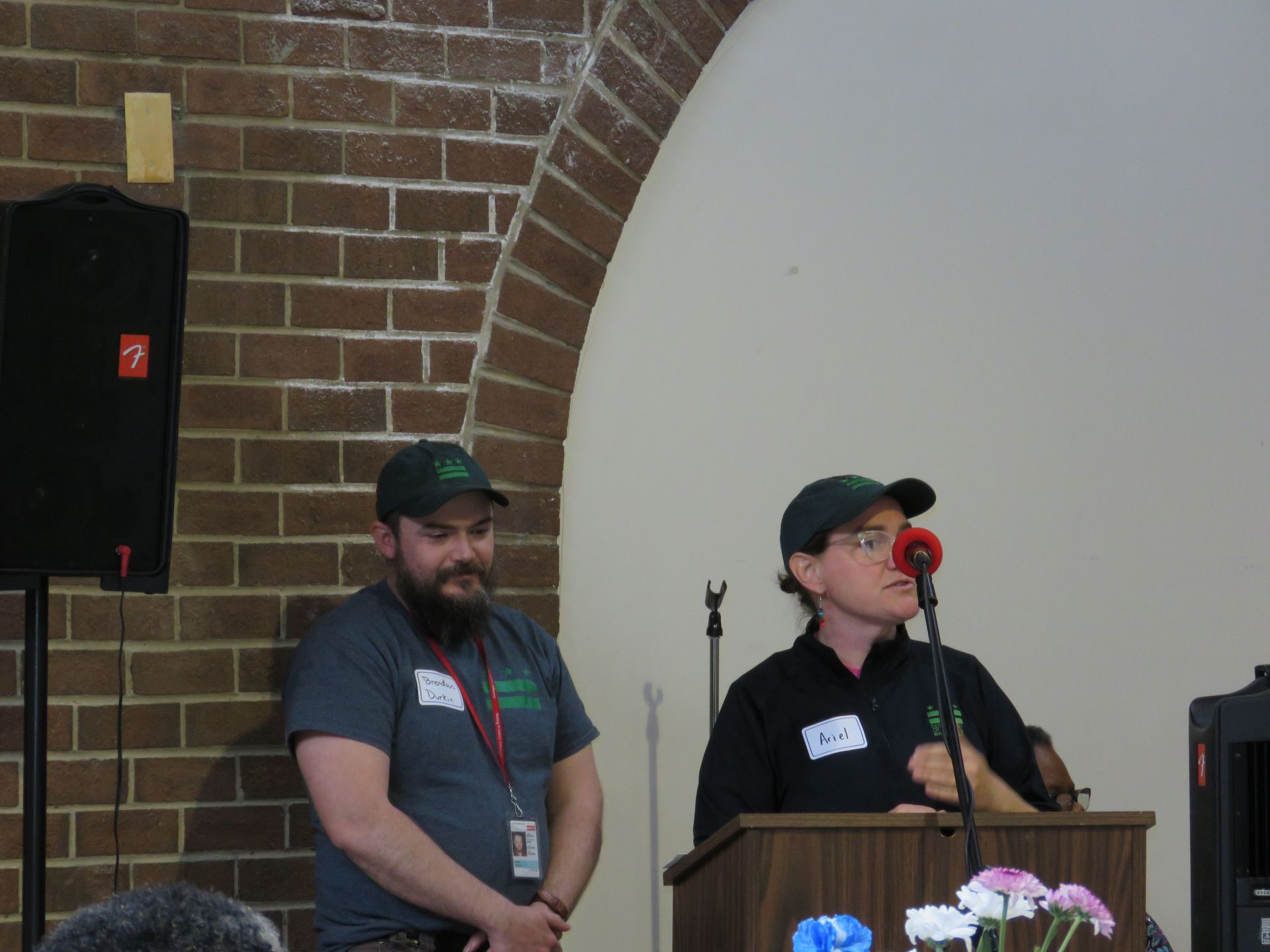 Two people in matching green caps and shirts on stage at a podium; one speaking into microphone, other standing.