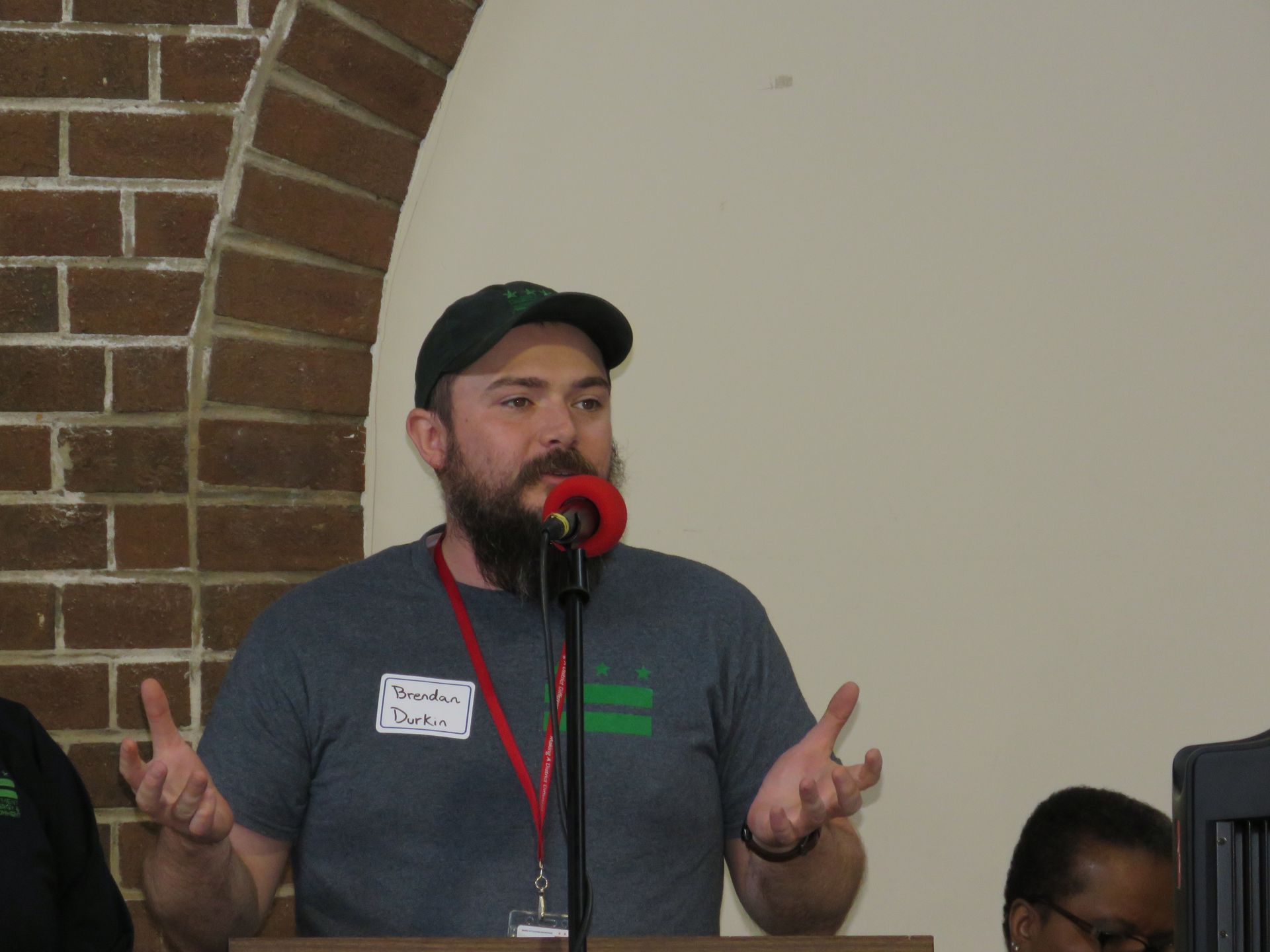 Man with beard speaking at podium, gesturing with hands. Brick wall background.