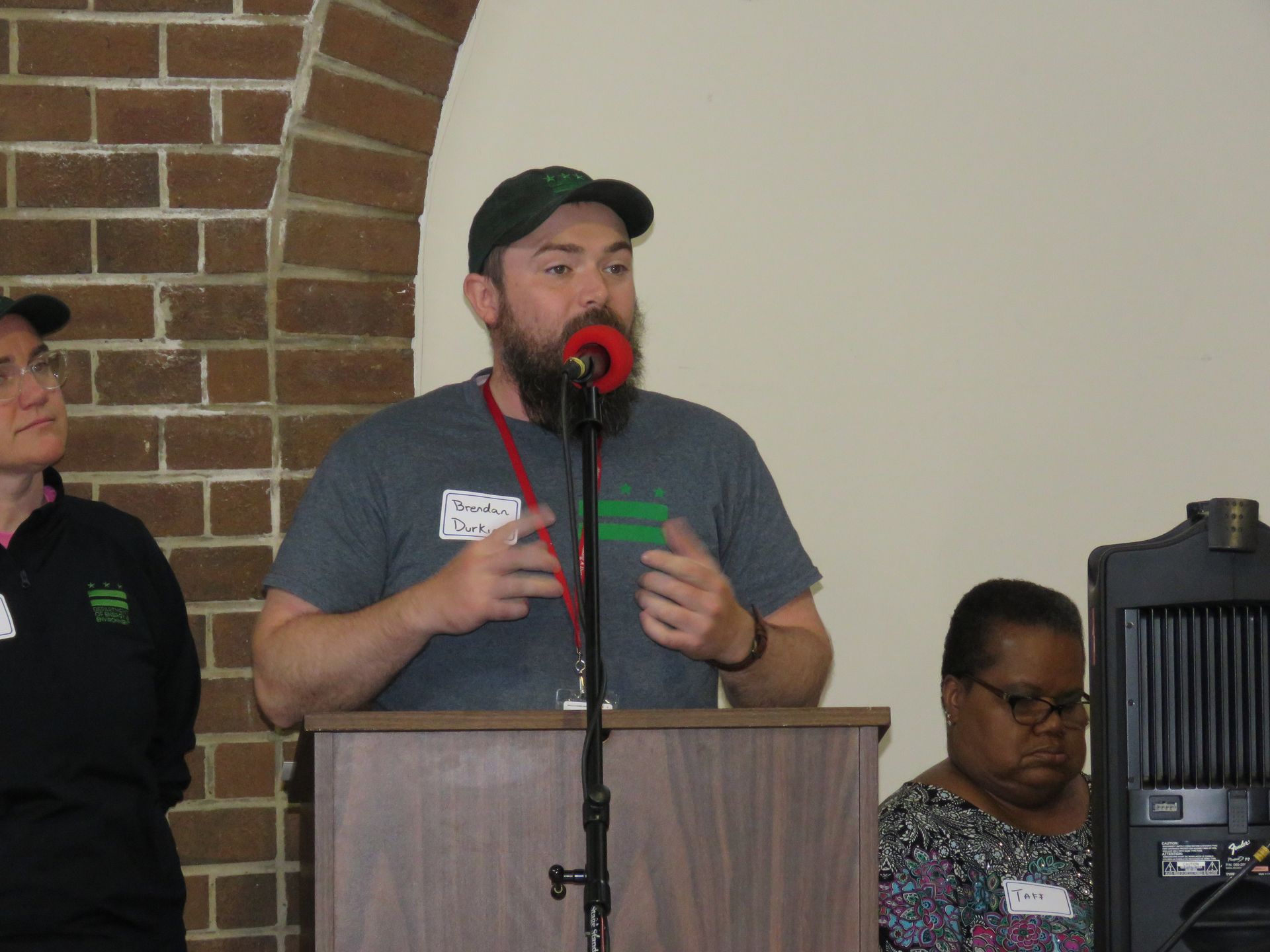 Man speaking at a podium, wearing a green hat and grey shirt. Two people beside him, brick arch background.