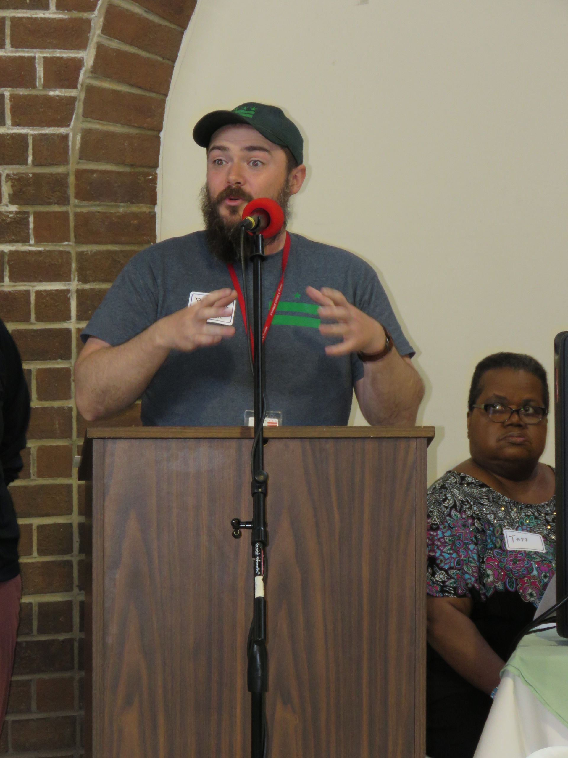 Man speaking at a podium, gesturing with hands. A woman sits to the right, looking on. Brick and beige background.