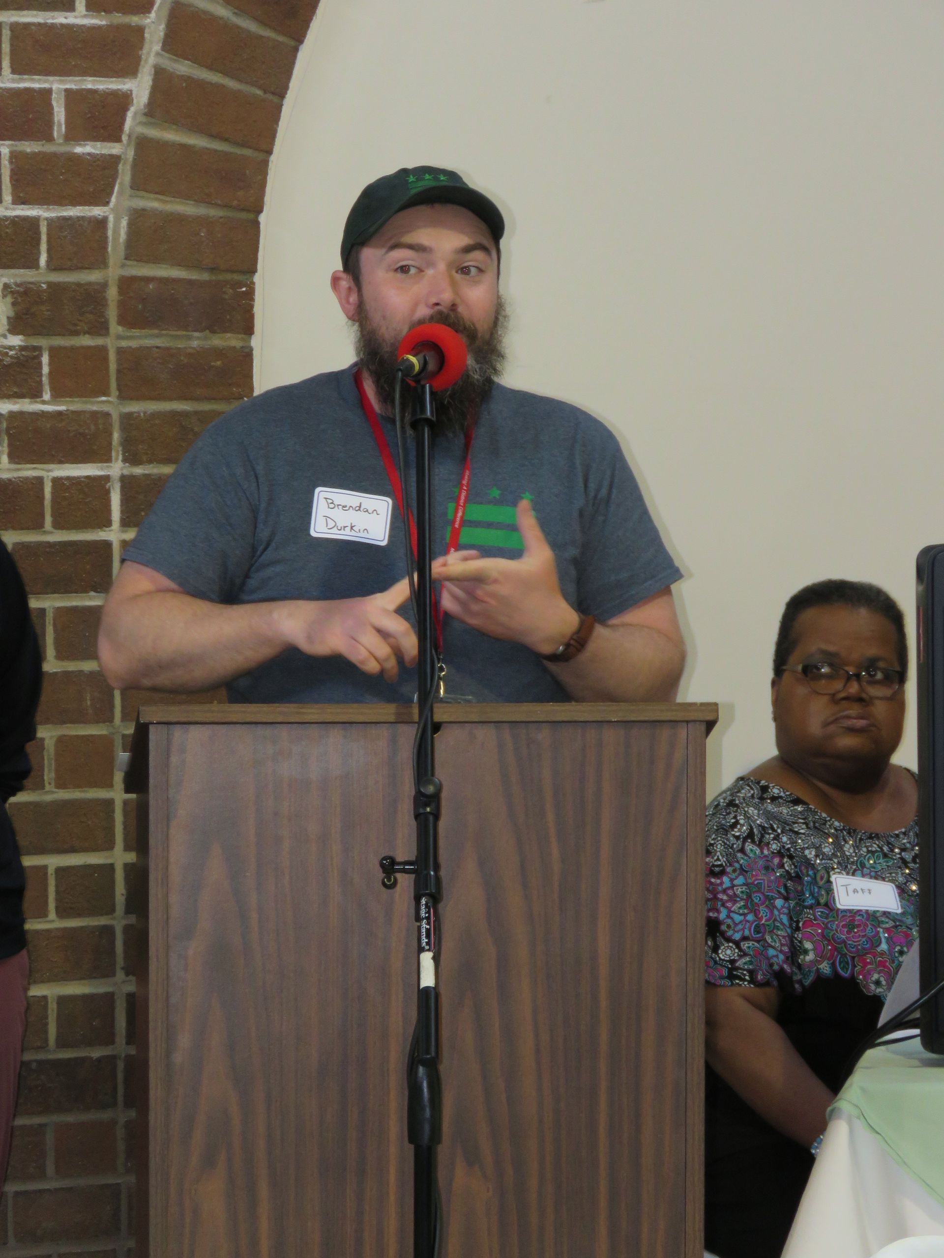 Man with beard speaking at a podium with microphone. Woman sits nearby, observing. Brick wall and white background.