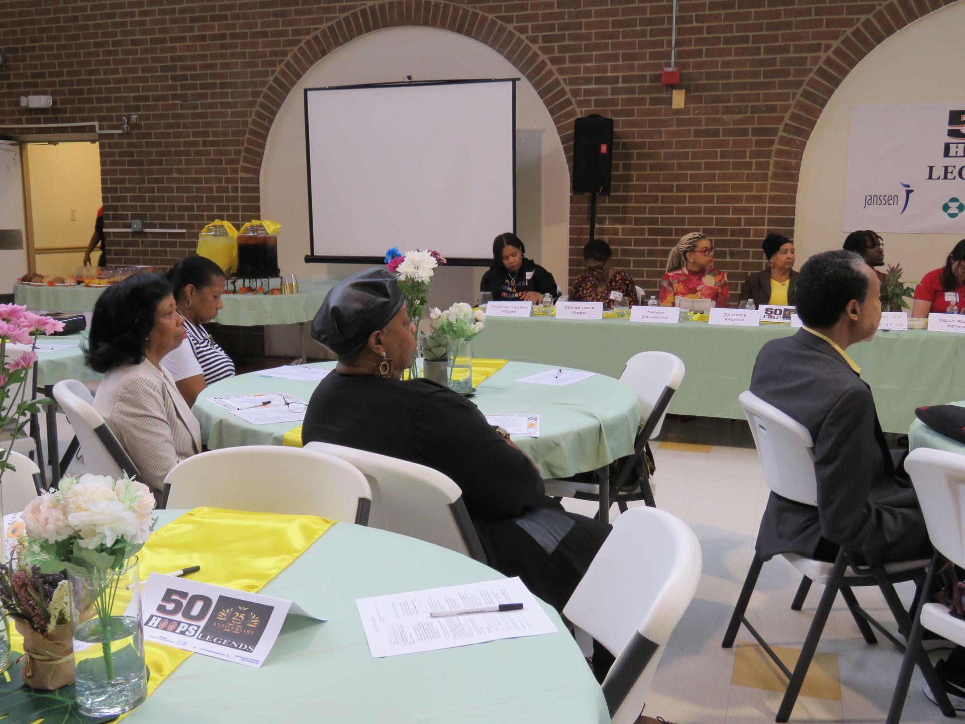 People seated at tables in a room, watching a panel at a presentation with a screen and brick walls.