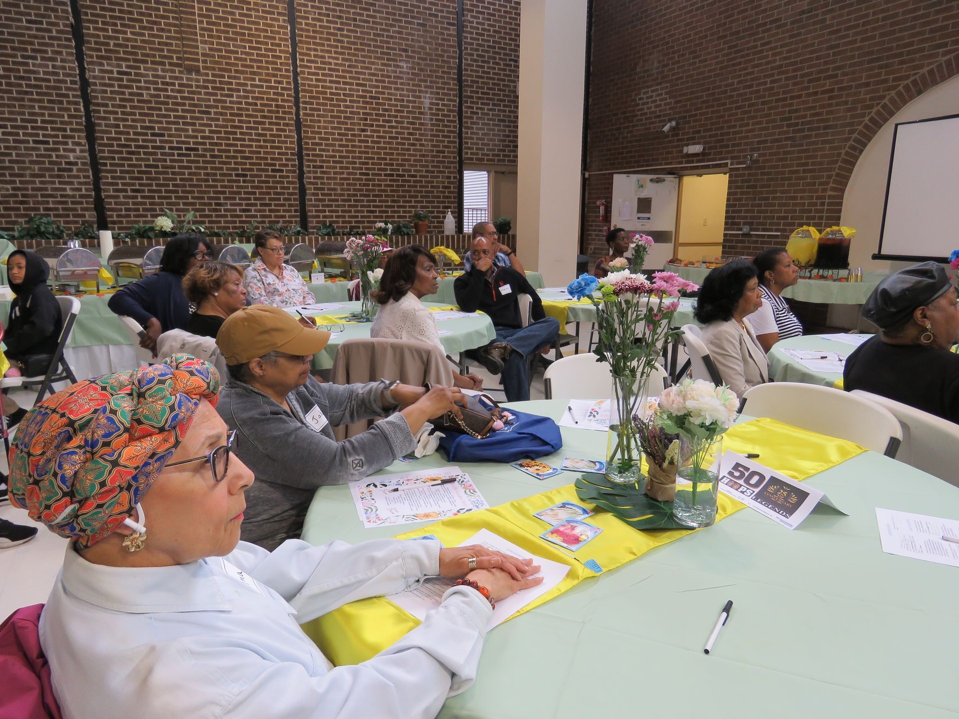 People seated at tables, attending an event in a hall with brick walls and floral centerpieces.
