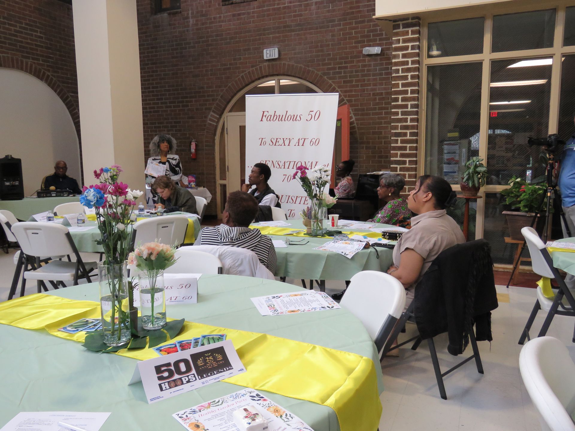 People seated at tables during an event. Tables are decorated with flowers and centerpieces.