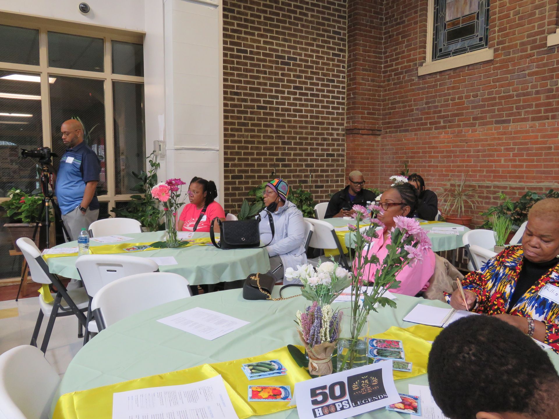 People seated at tables during an event, some with pens. A person films the event. Indoor setting with brick wall.