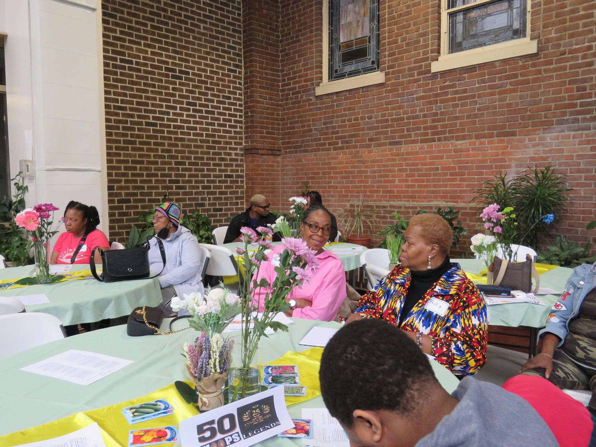 People seated at tables decorated with flowers, outdoors near a brick building.