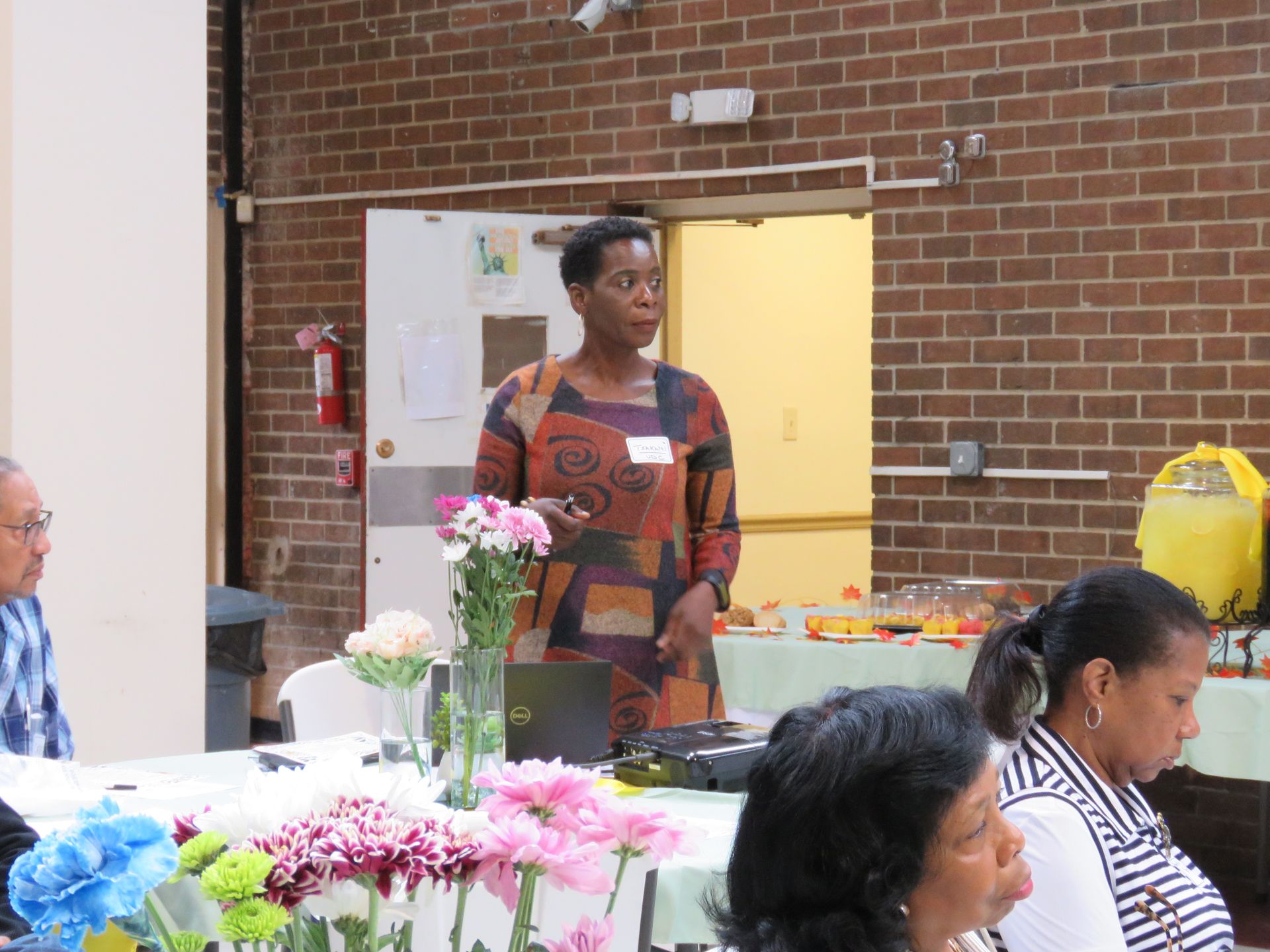 Woman speaking at a table, flowers in foreground, brick wall backdrop, people seated.