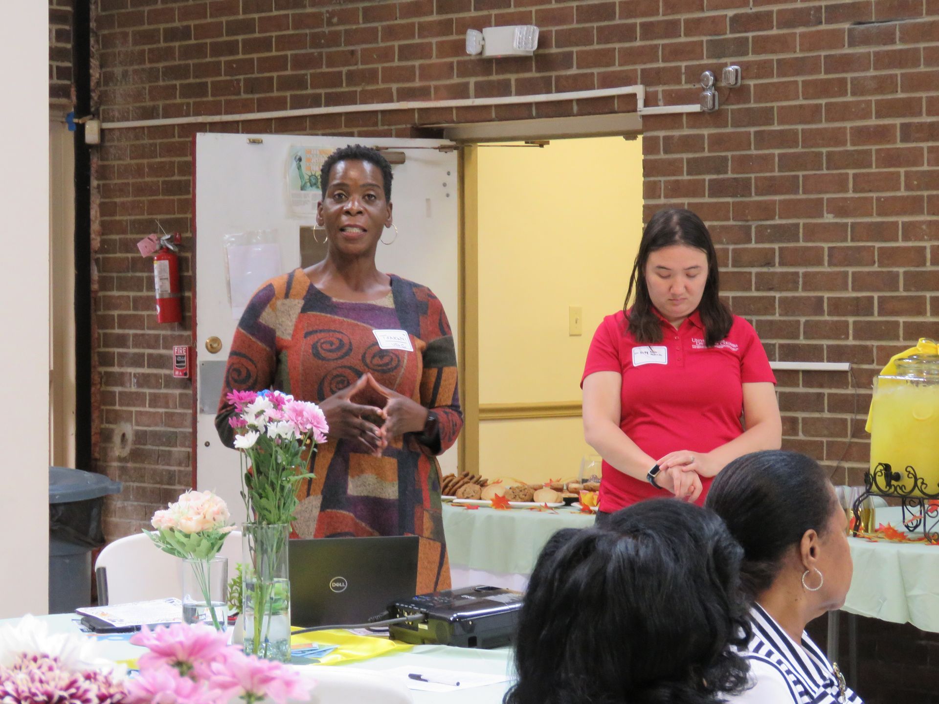 Woman speaking at a table with a laptop, another woman standing nearby, and audience in a room.