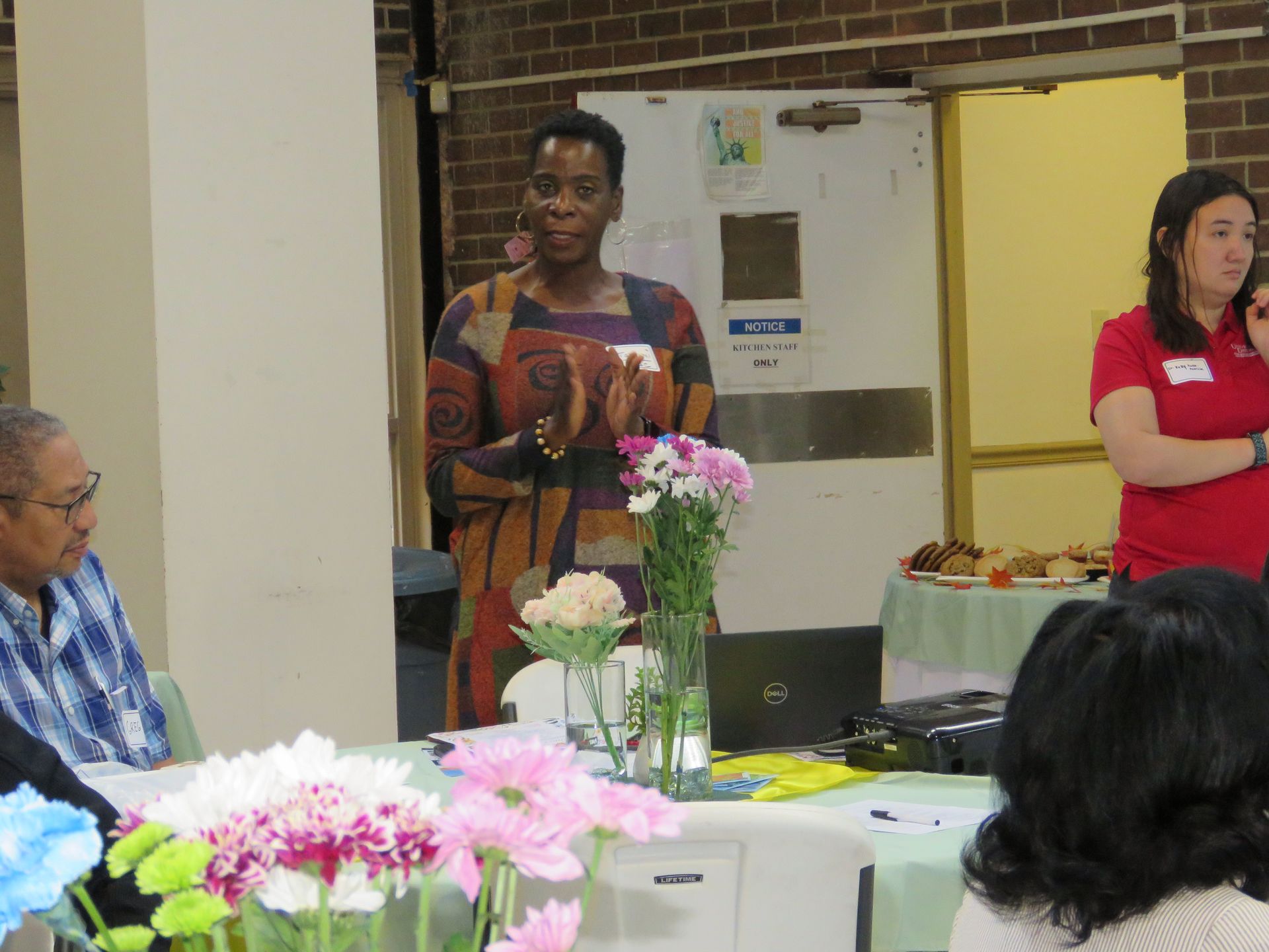 Woman in patterned dress speaking at table with flowers, laptop, and snacks. Another woman stands to the side.