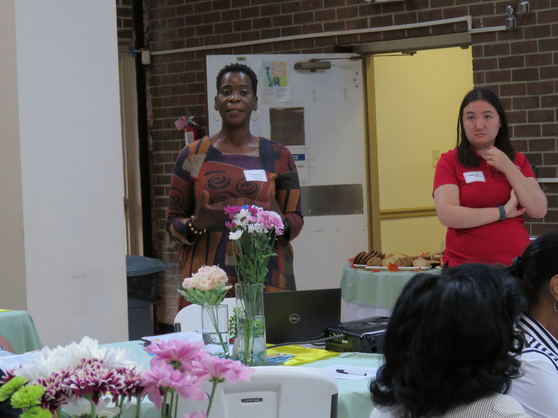 Woman speaking at a table with flowers, another woman stands to the side. Both wear name tags, interior setting.