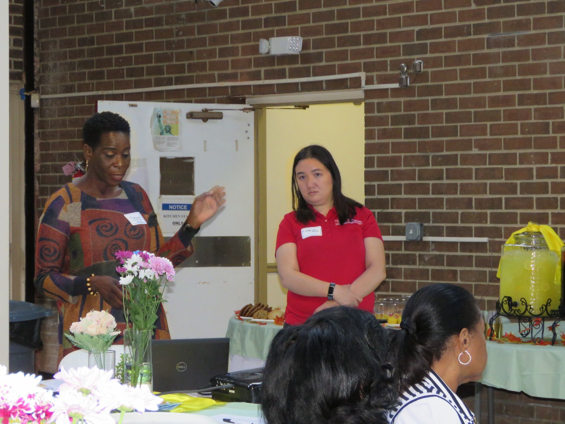 Two women at a presentation in a room, one speaking, gesturing. Attendees sit at a table.