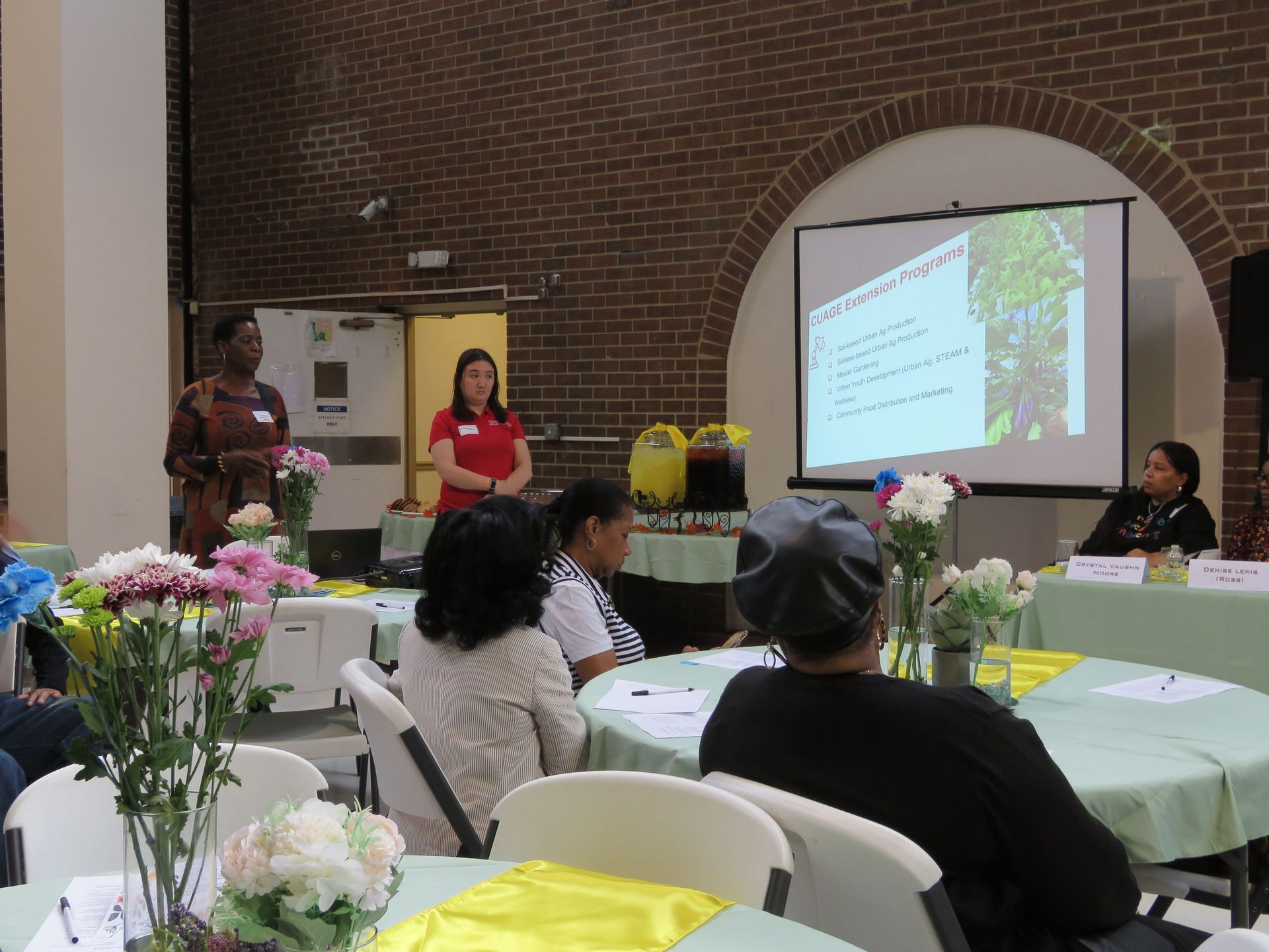 People at a presentation: two speakers, attendees seated at tables, a projection screen, and floral centerpieces.