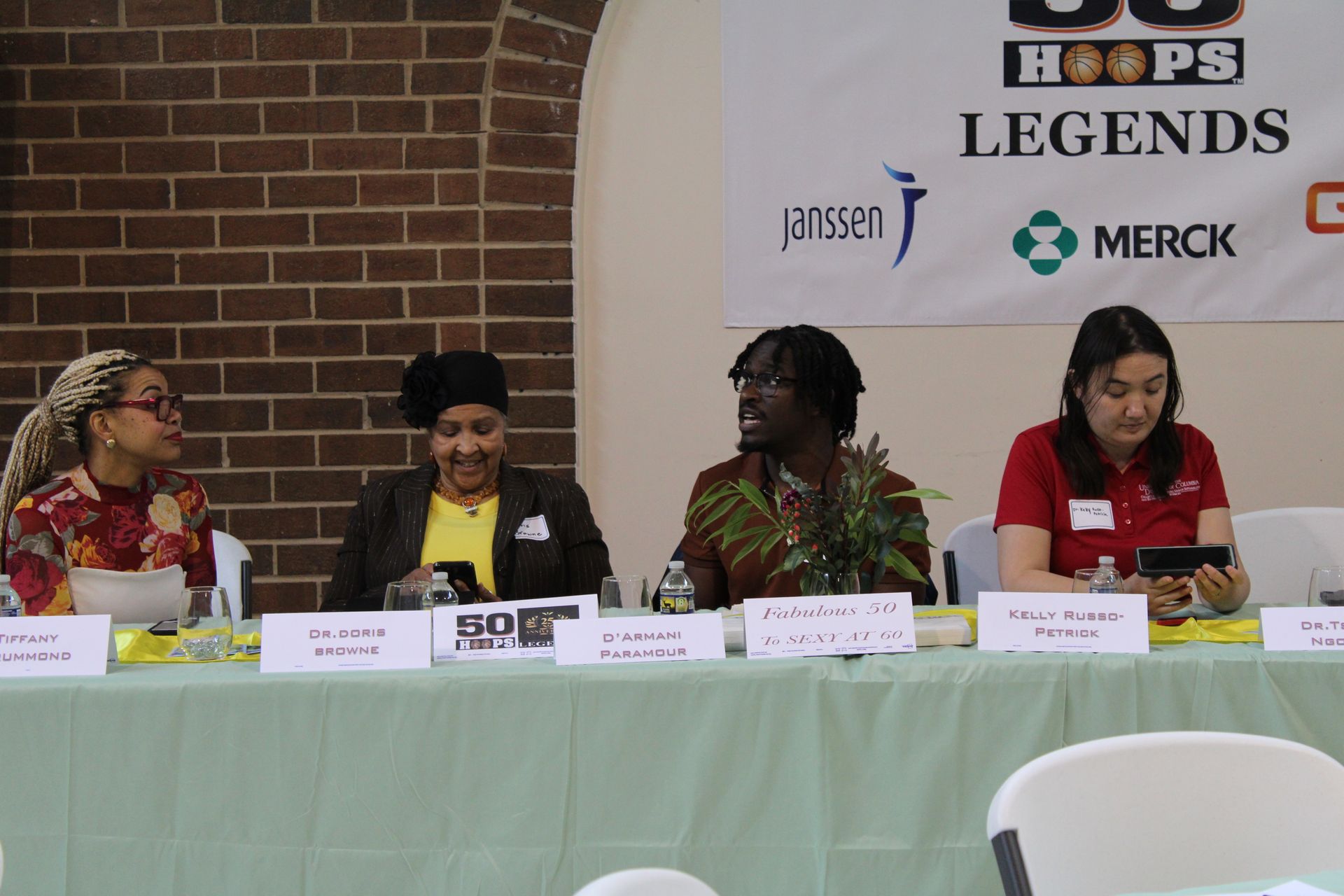 Panel of four speakers at a table with a banner that has sponsors' logos and text.