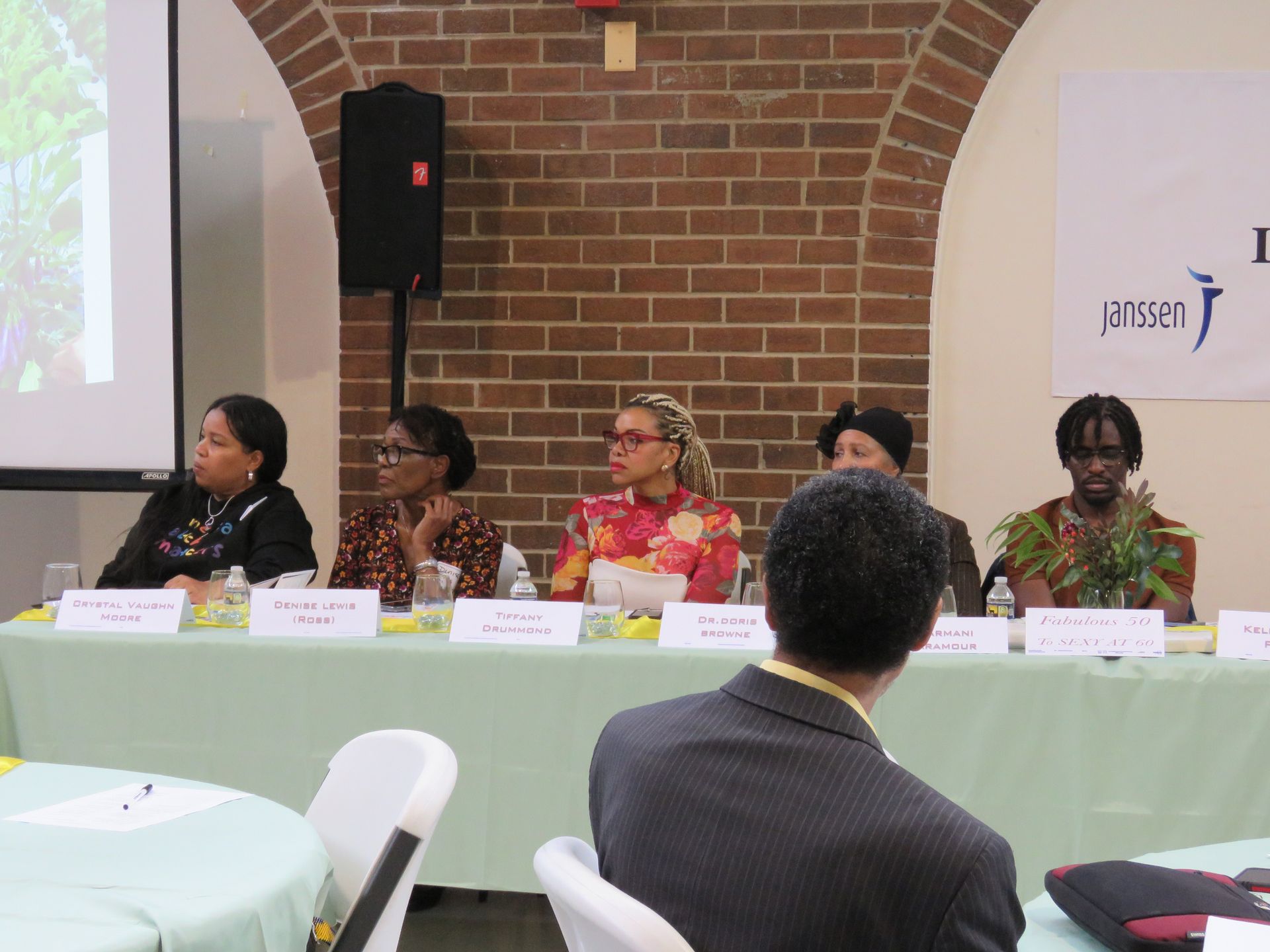 Panel of people at a conference, sitting behind a table, speaking to the audience. Brick wall in the background.