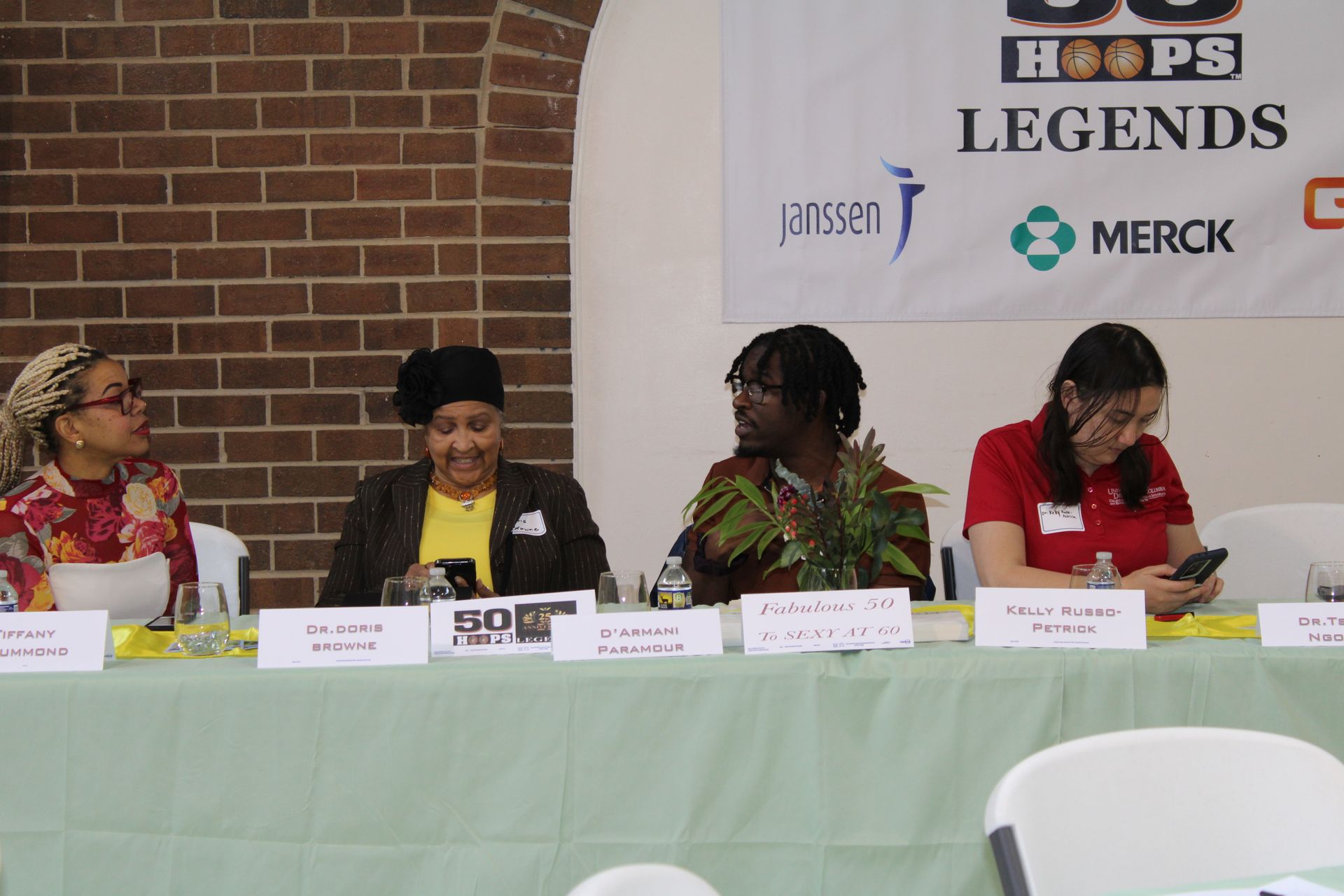 Panel of four people seated at a table with a banner that has sponsors.